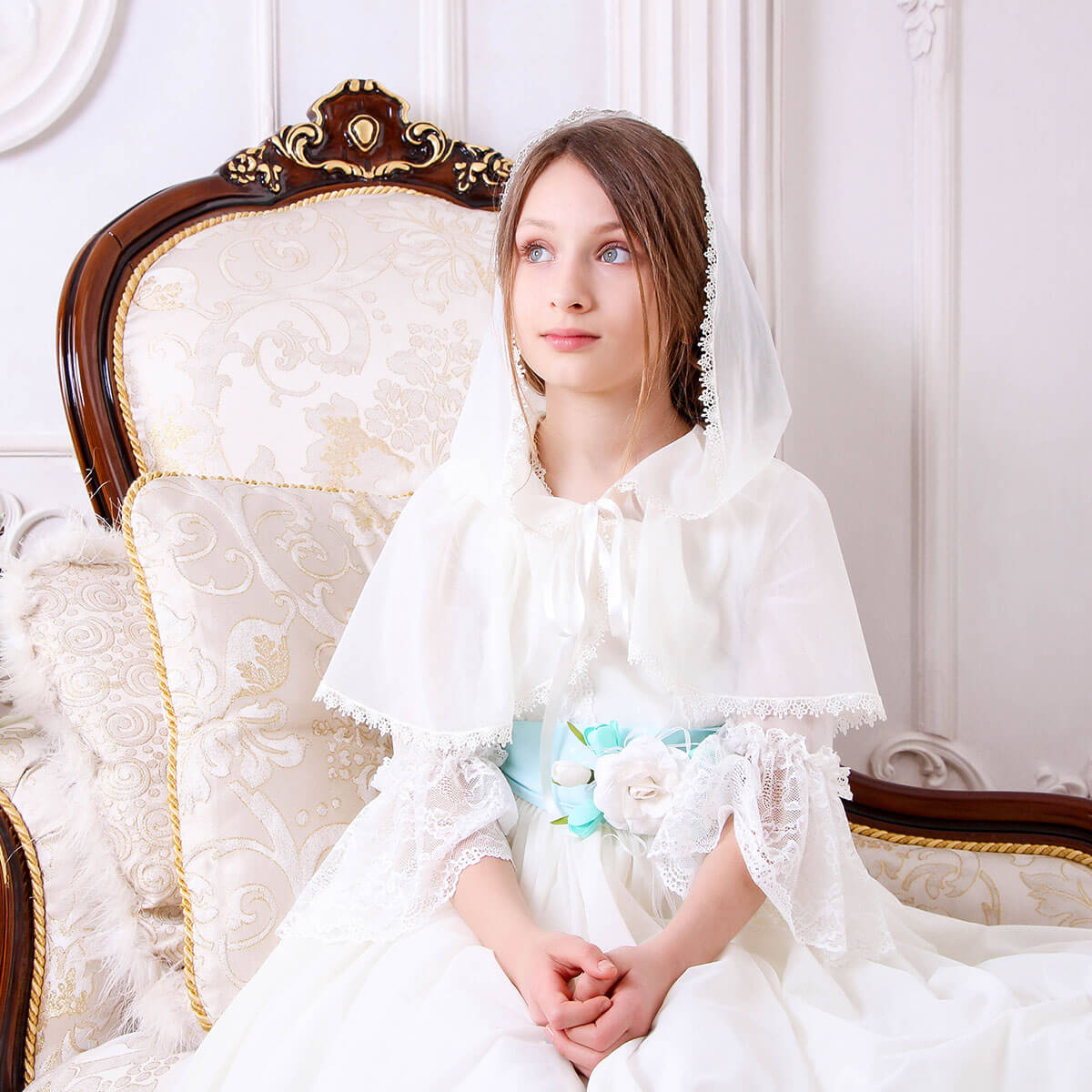 Young girl in a Traditional Communion Dress with a floral sash sitting on an ornate chair.