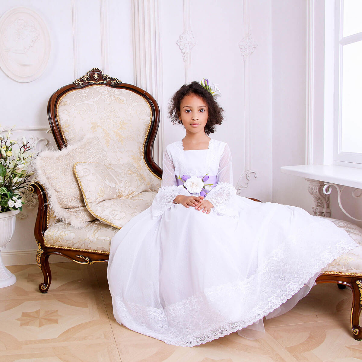 Young girl in a Traditional Communion Dress sitting on an ornate chair in a bright room.