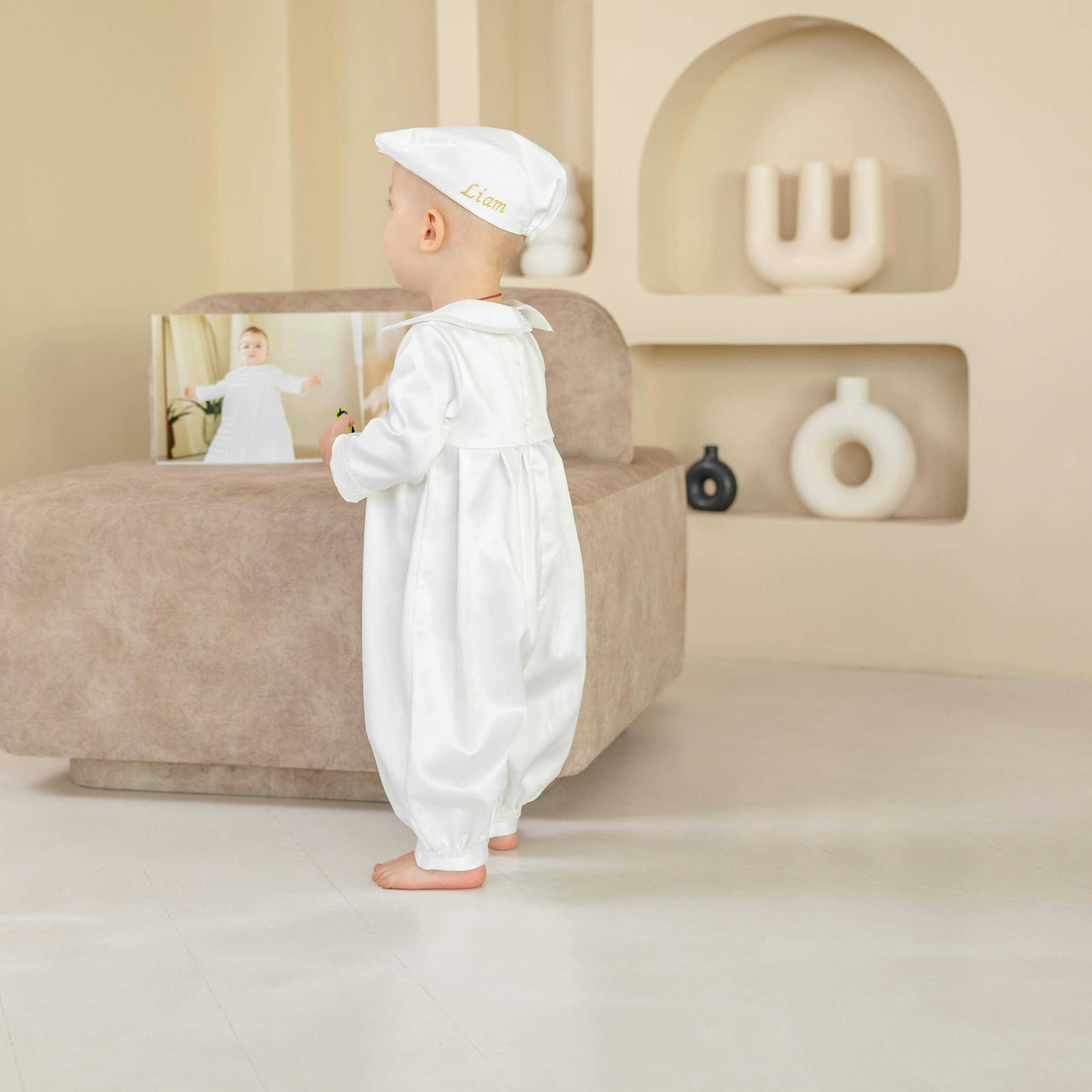 Baby in a  Baby Boy Christening Romper standing in a minimalistic room with a beige sofa and decorative items.