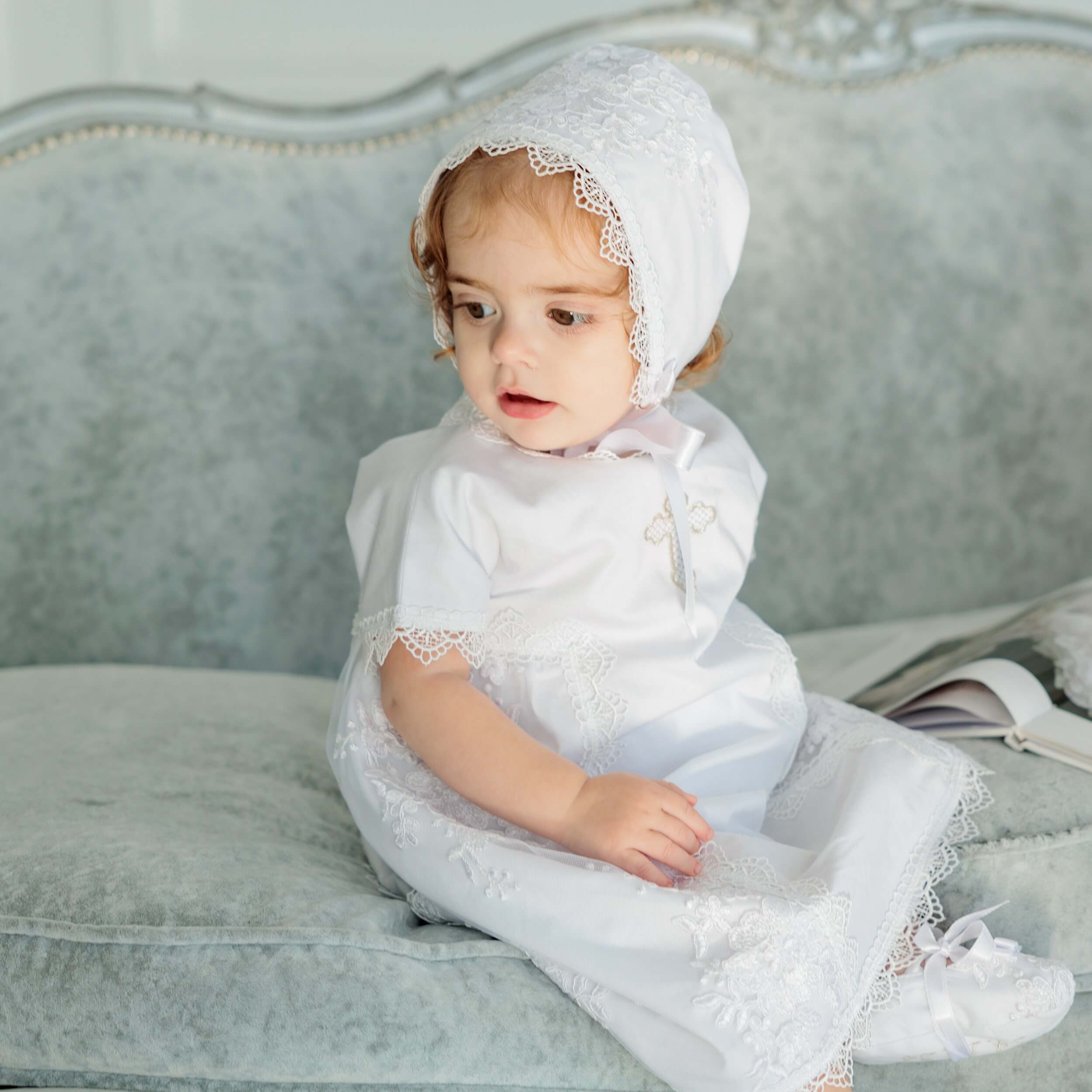 Baby in lace bptism bonnet n a gray couch.