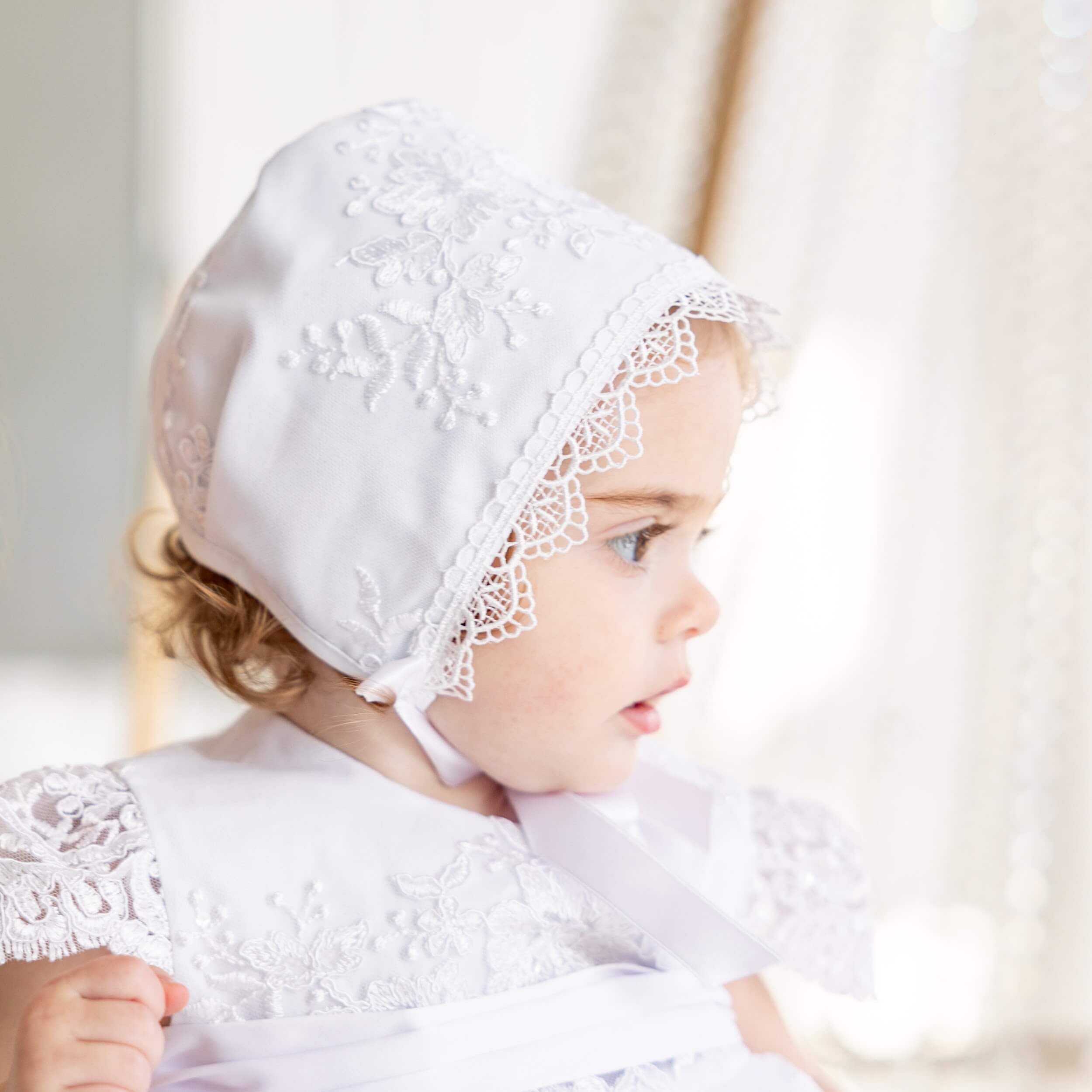 Baby wearing lace bptism bonnet and dress against a neutral background