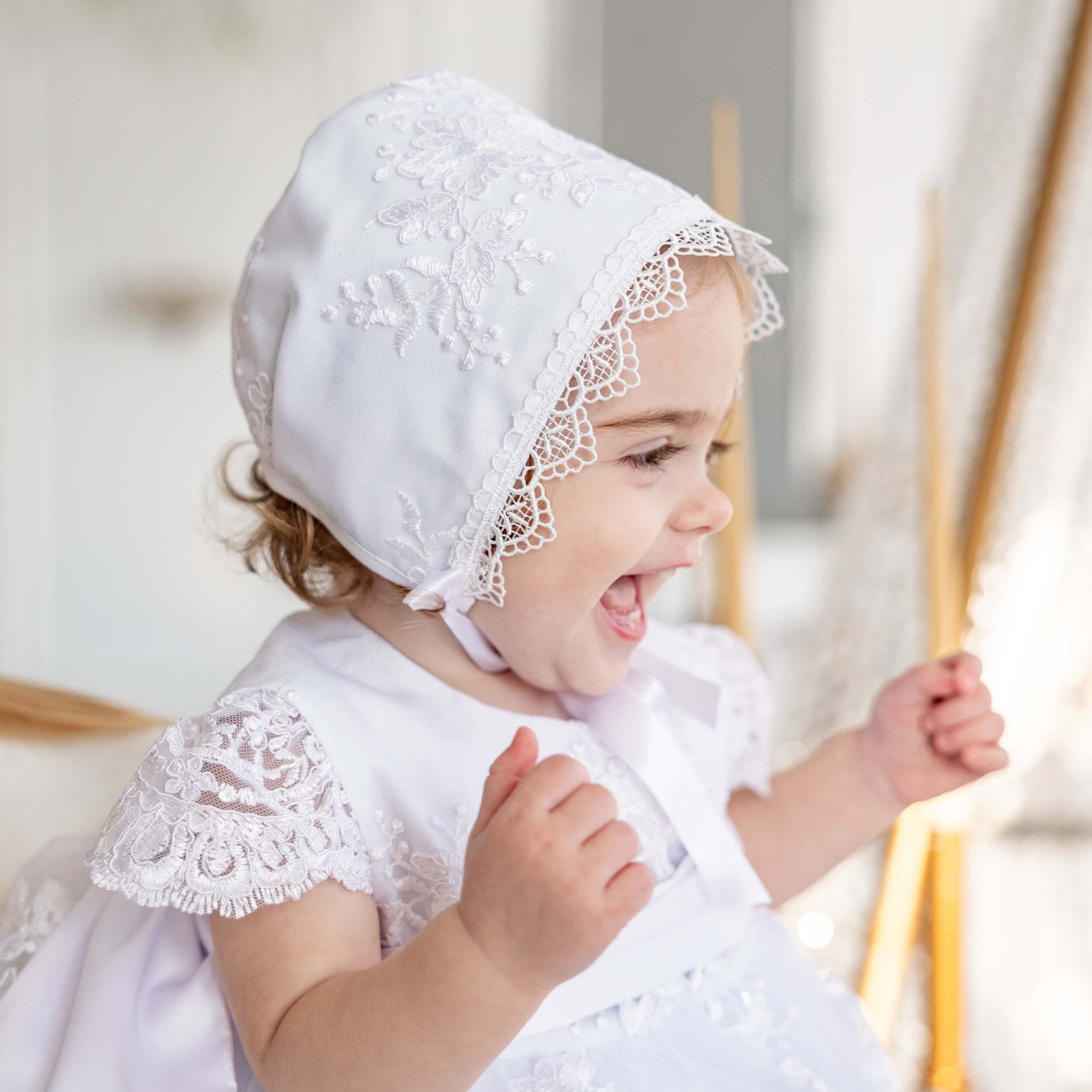 Child wearing Lace Baptism Bonnet and dress, smiling in a softly blurred indoor setting.