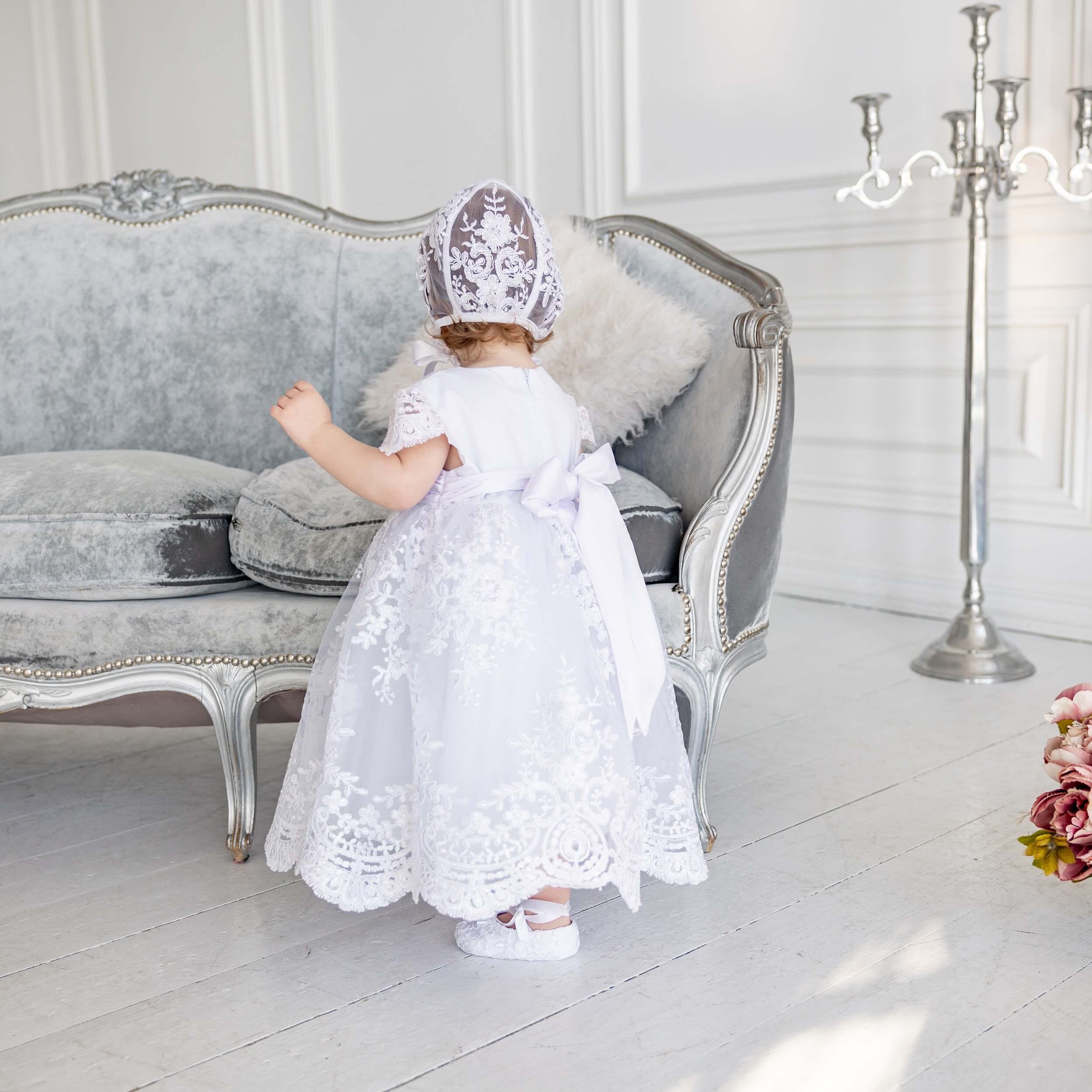 Child in a Lace Baptism Dress and bonnet standing in a elegant room with a sofa and candelabra.