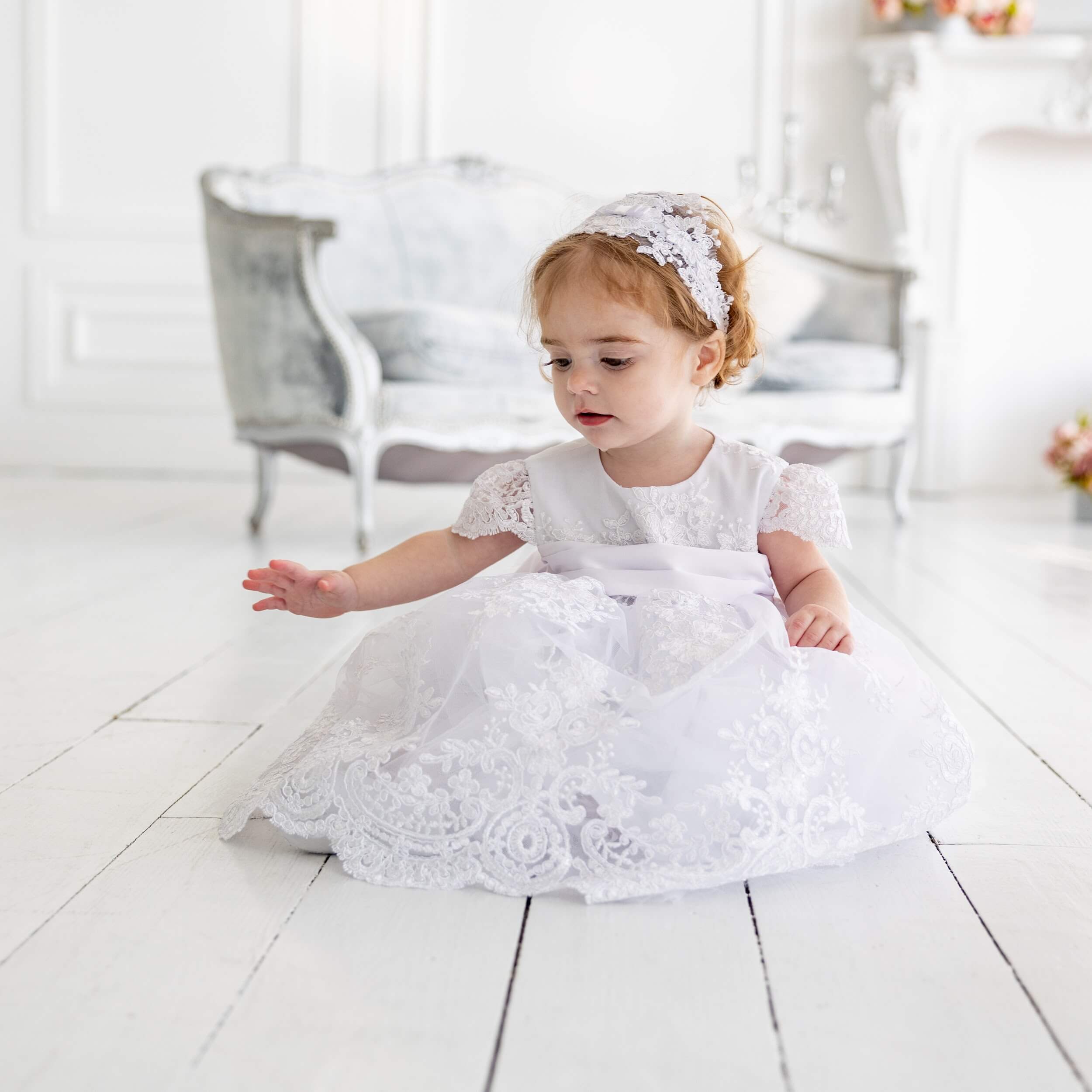 Child in a Lace Baptism Dress sitting on a light wooden floor.