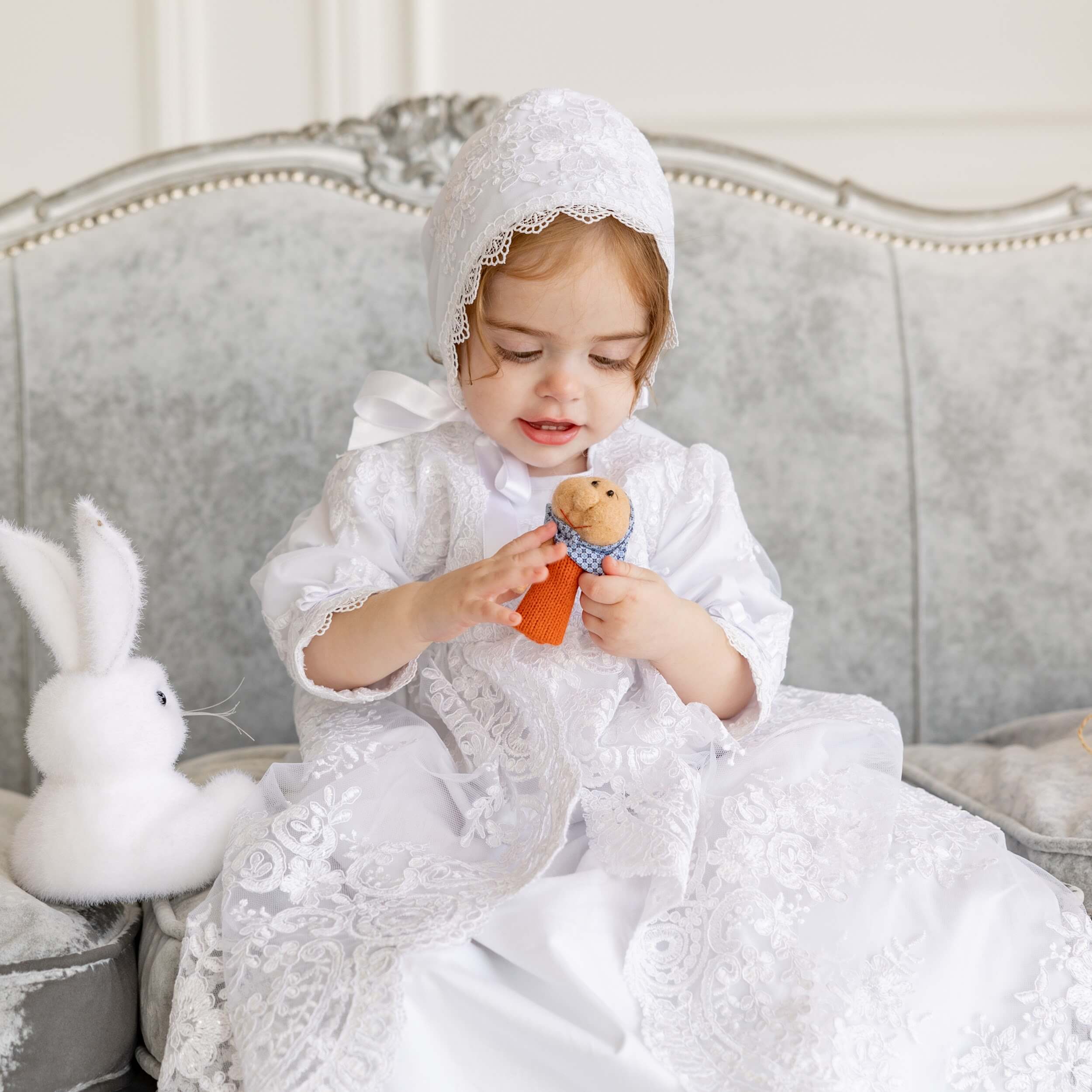 Child in a Christening Gown for Girls sitting on a gray sofa with a teddy bear.