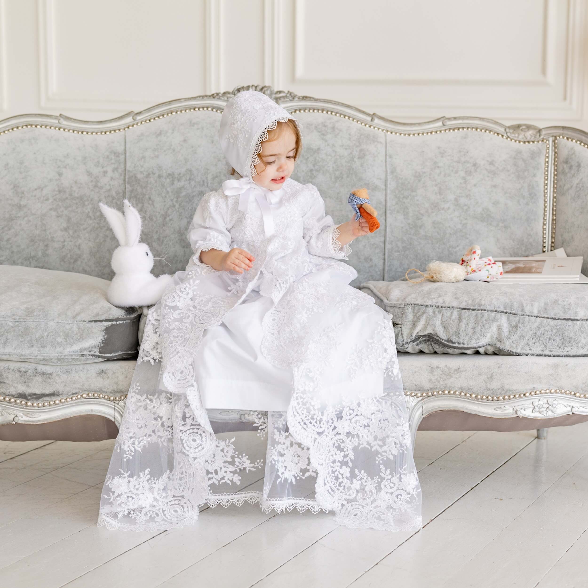 Child in a Christening Gown for Girls sitting on a gray sofa with toys.