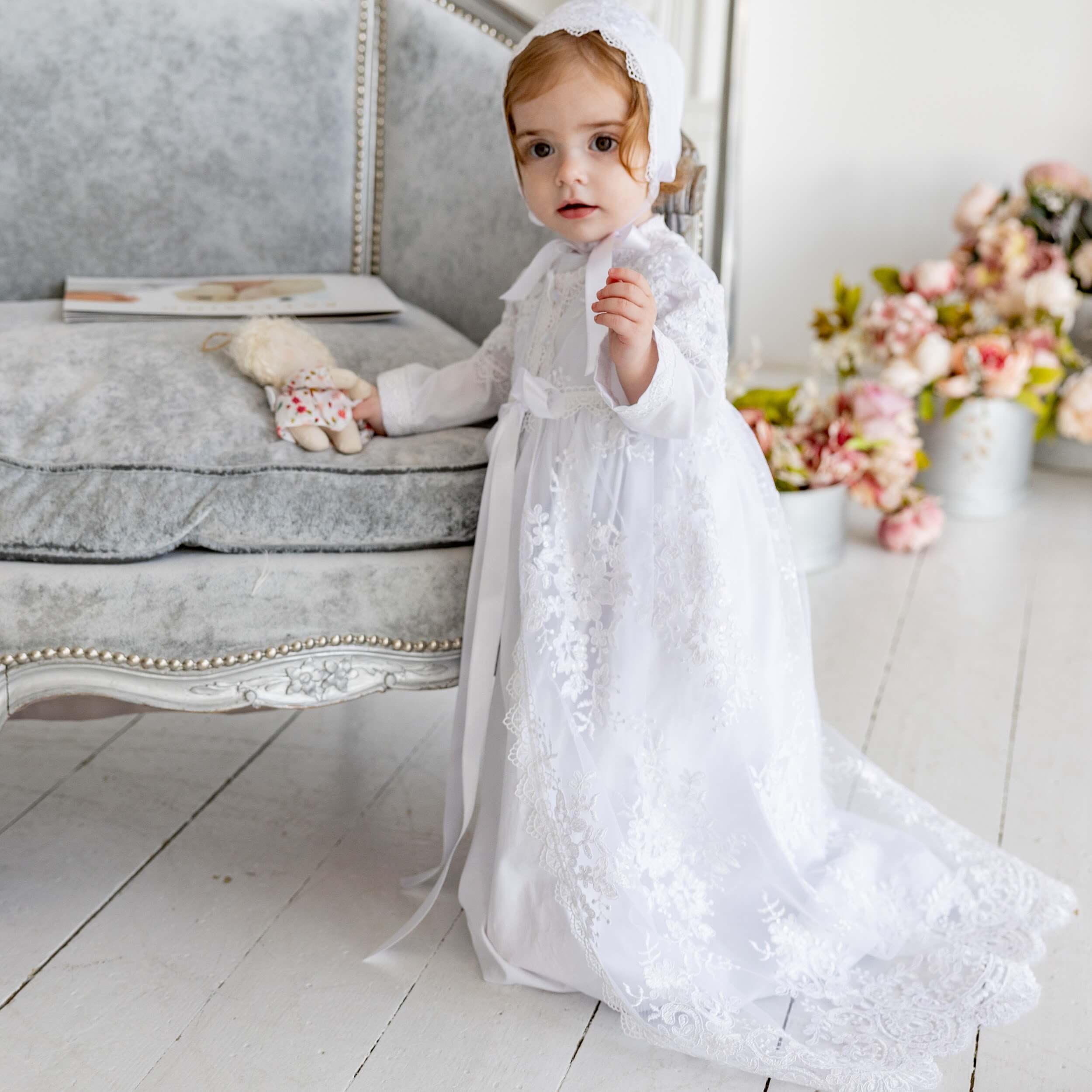 Child in a  Christening Gown for Girls  sitting on a light wooden floor with a floral arrangement in the background.
