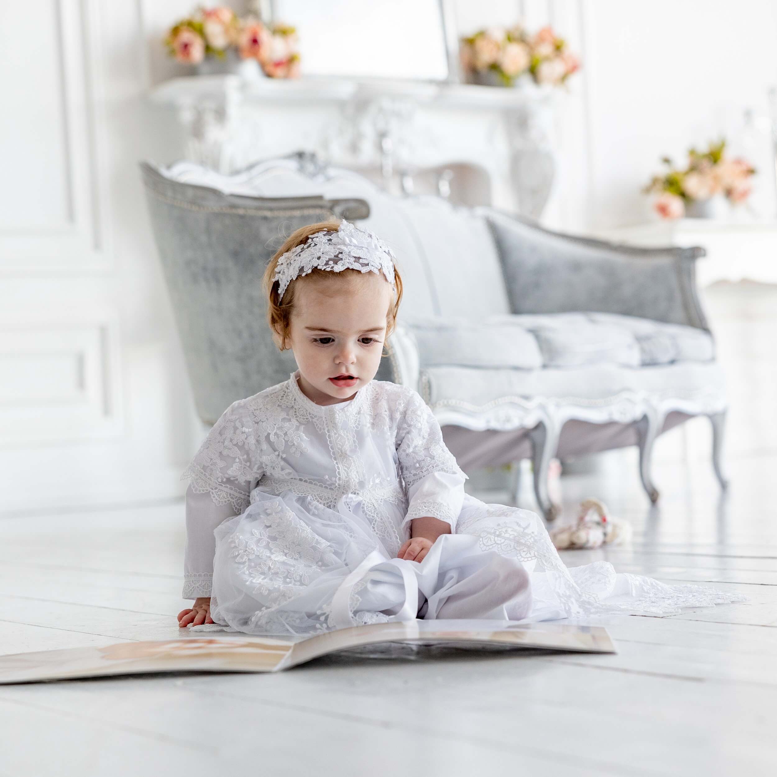 Child in a  Christening Gown for Girls  sitting on a light wooden floor with a book open in front of them.