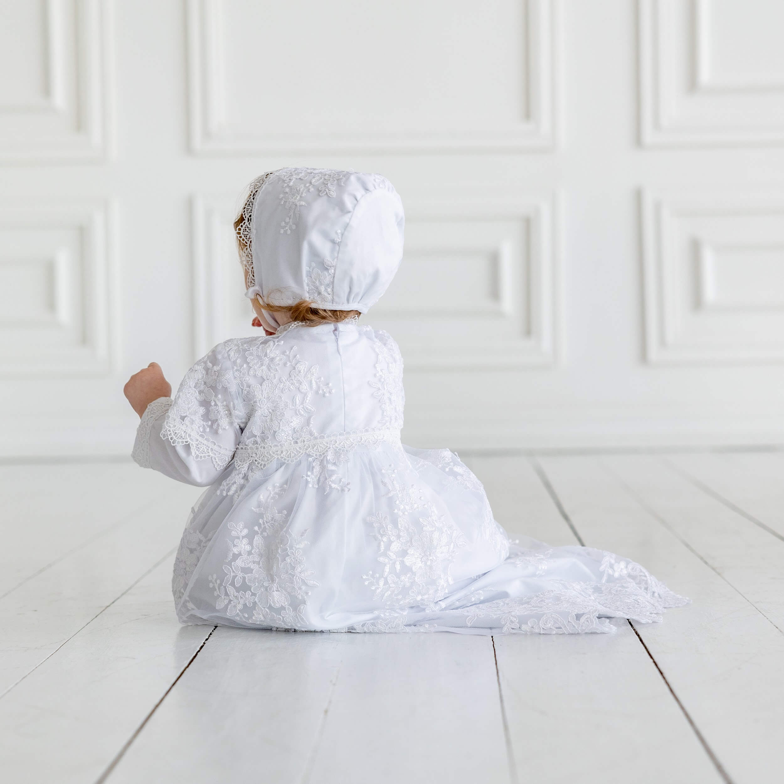 Child in a  Christening Gown for Girls and bonnet sitting on a white floor against a white paneled wall.