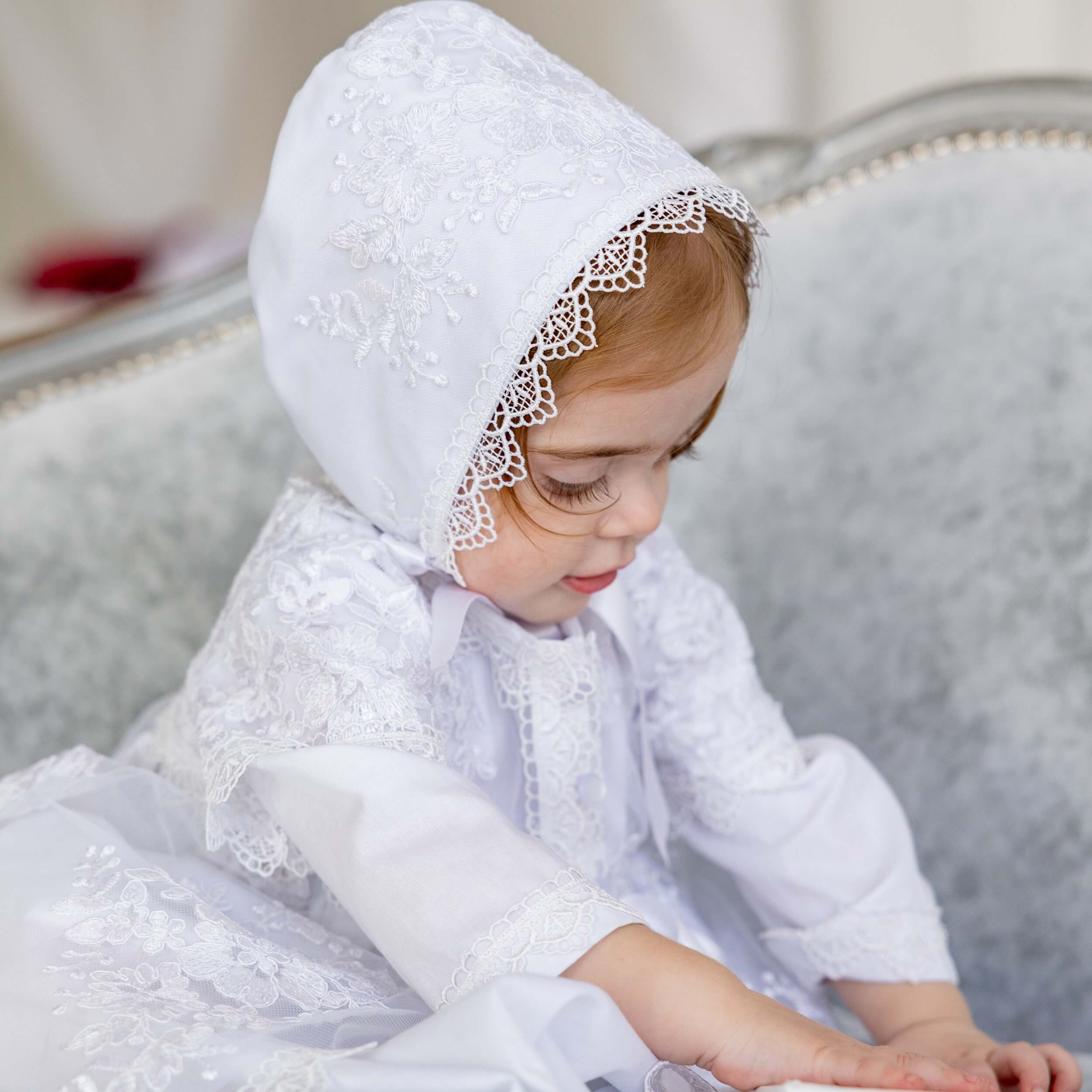 Child wearing a white lace bonnet and  Christening Gown for Girls  sitting on a couch.
