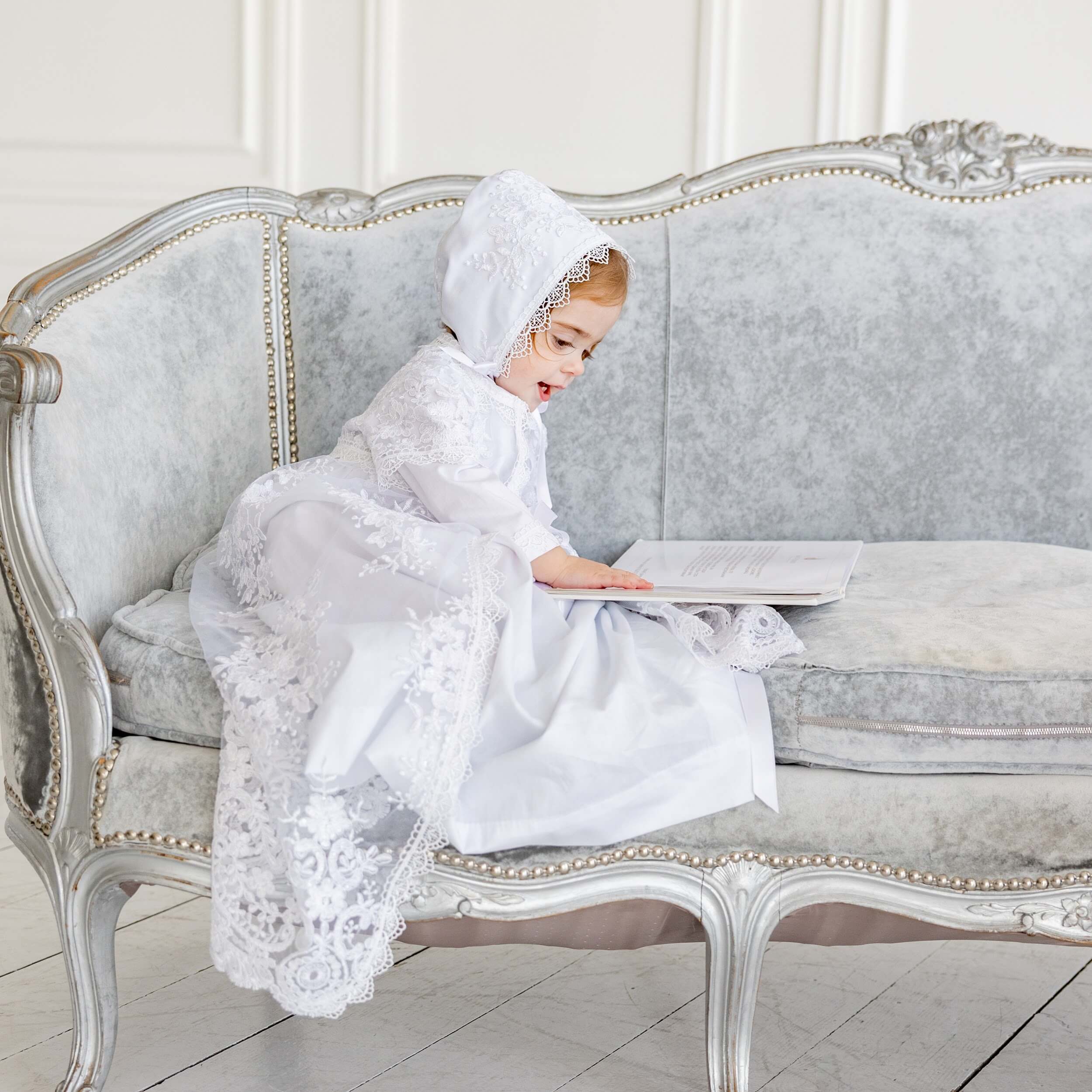 Child in a   Christening Gown for Girls  sitting on a decorative couch holding a book.