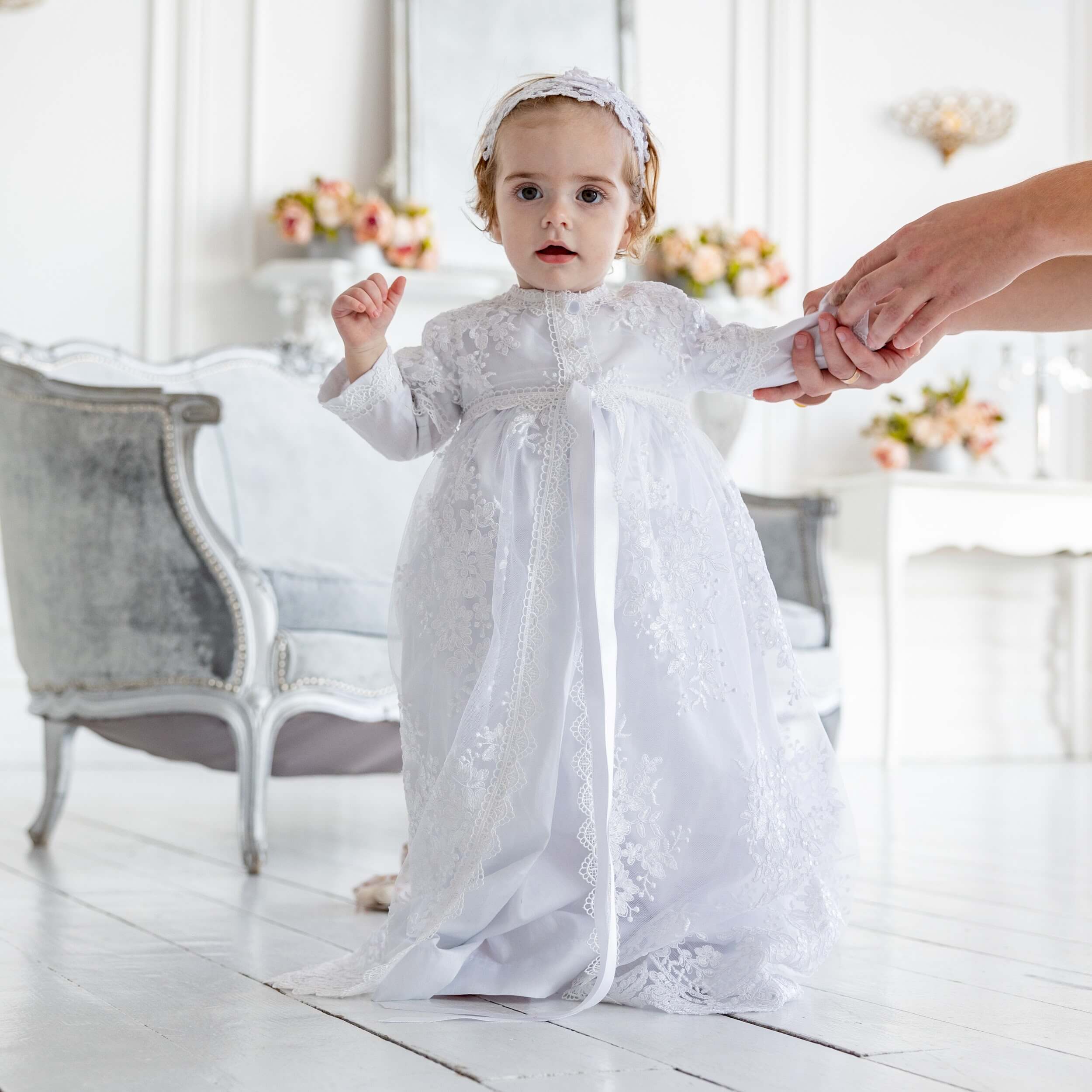 Child in a  Christening Gown for Girls standing on a light wooden floor with a blurred background
