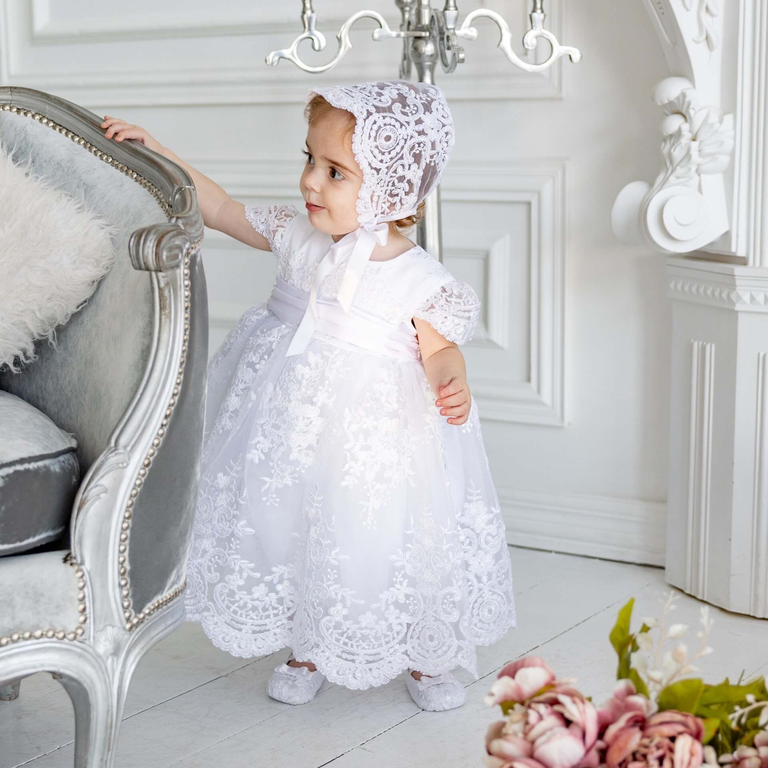 Young girl in a white lace dress and bonnet standing in a elegant room with floral decorations.