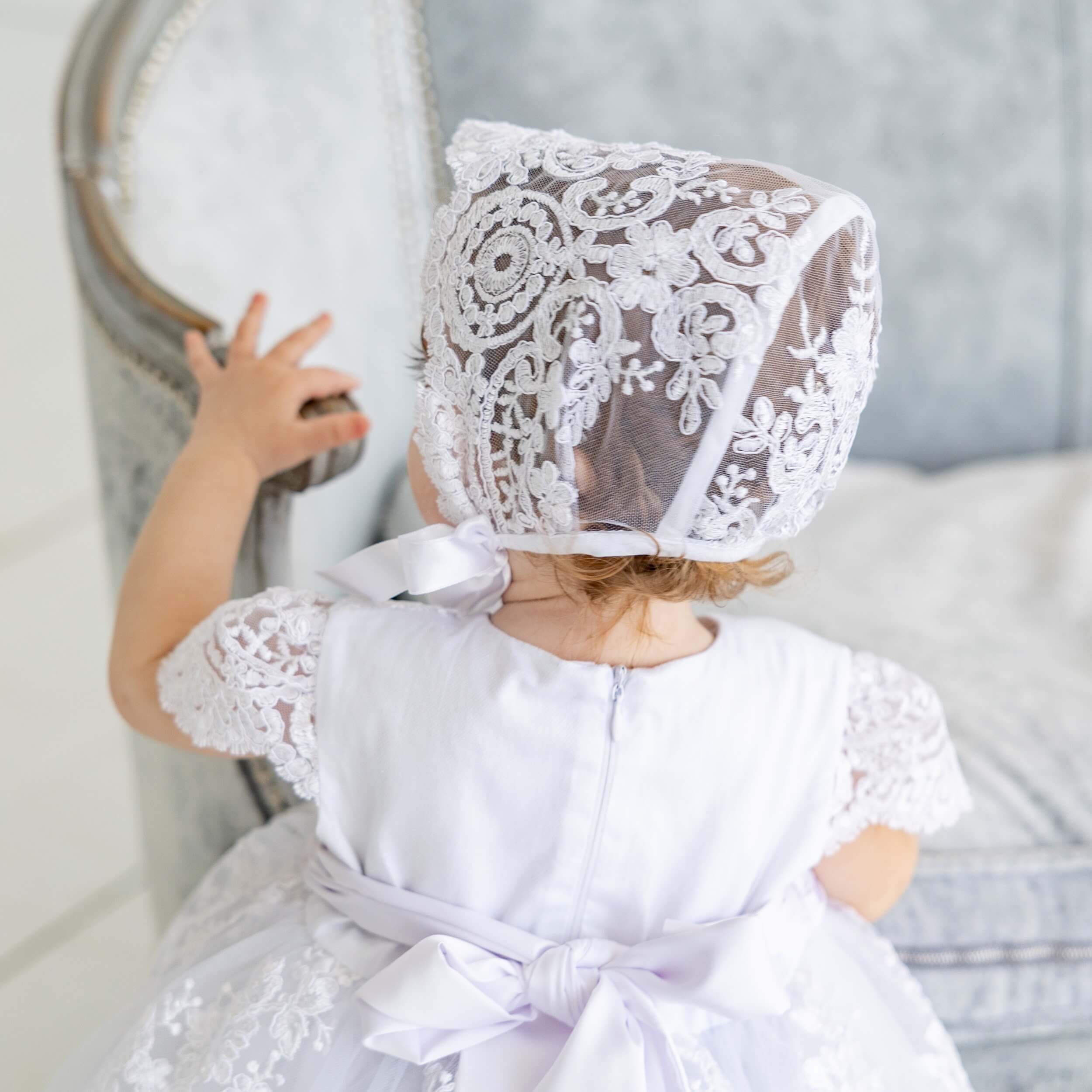 Child wearing a silk christening bonnet  and dress, standing in a neutral setting.
