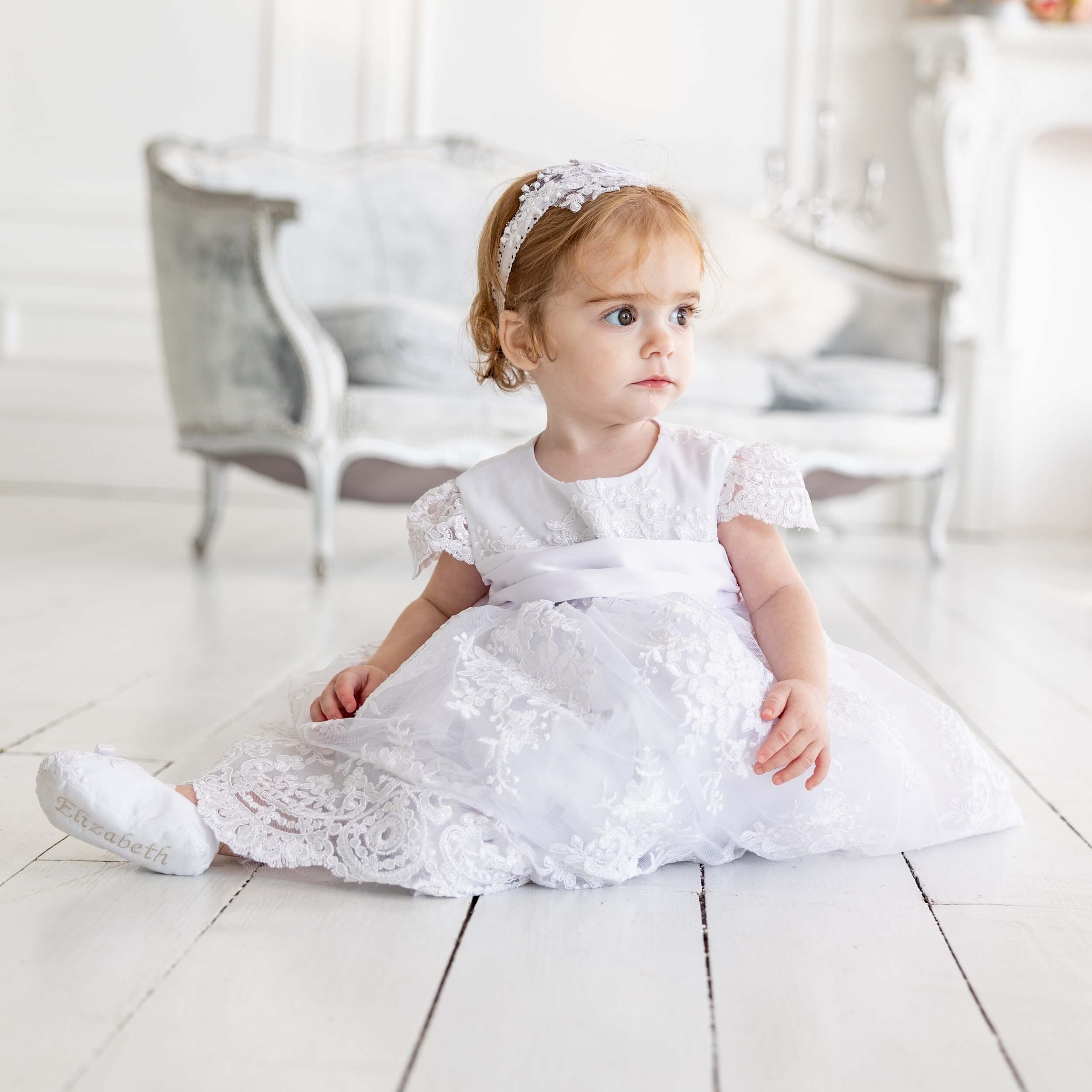 Young girl in a Baptism Dress for Girls sitting on a light wooden floor.