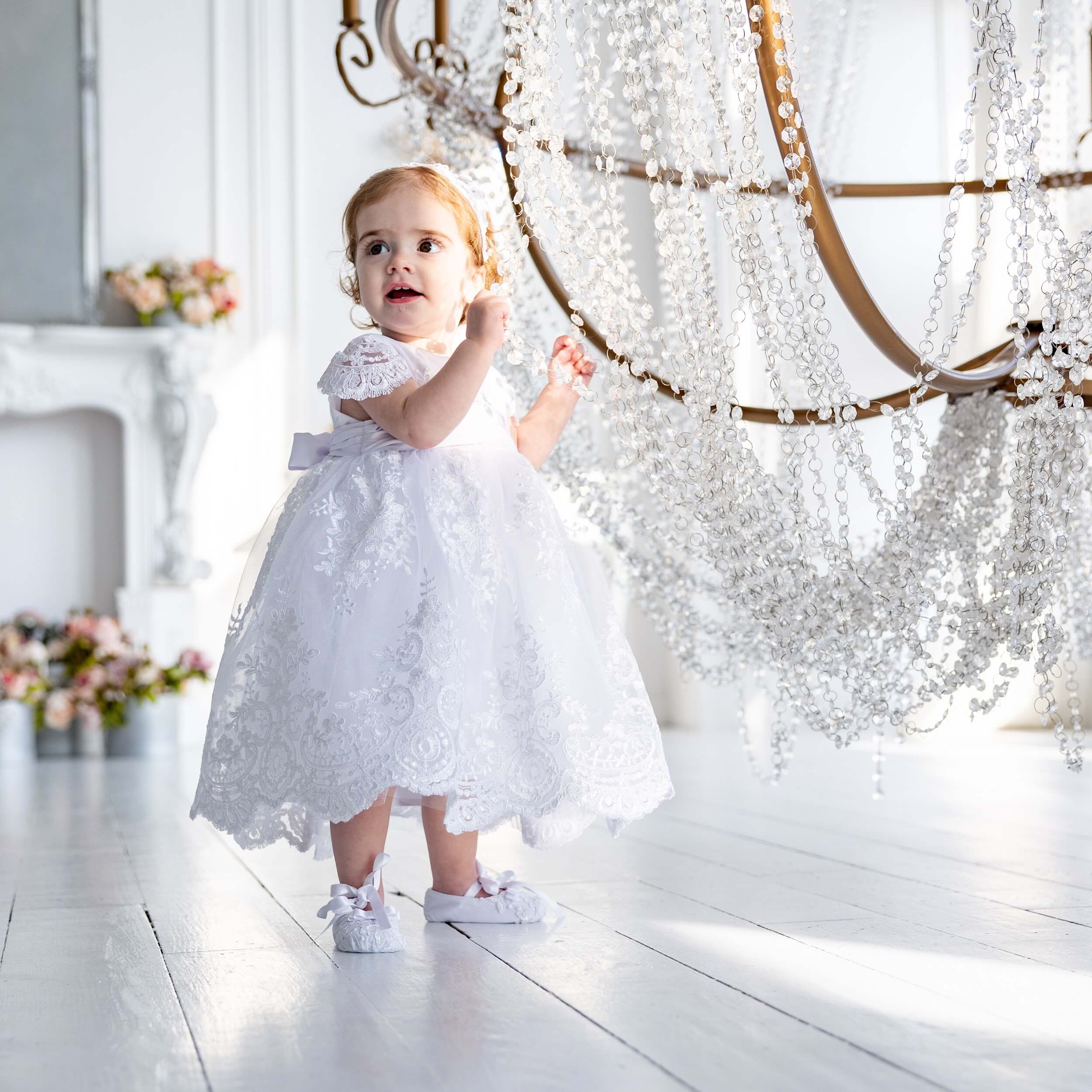 Child in a Baptism Dress for Girls standing in a bright room with a chandelier.