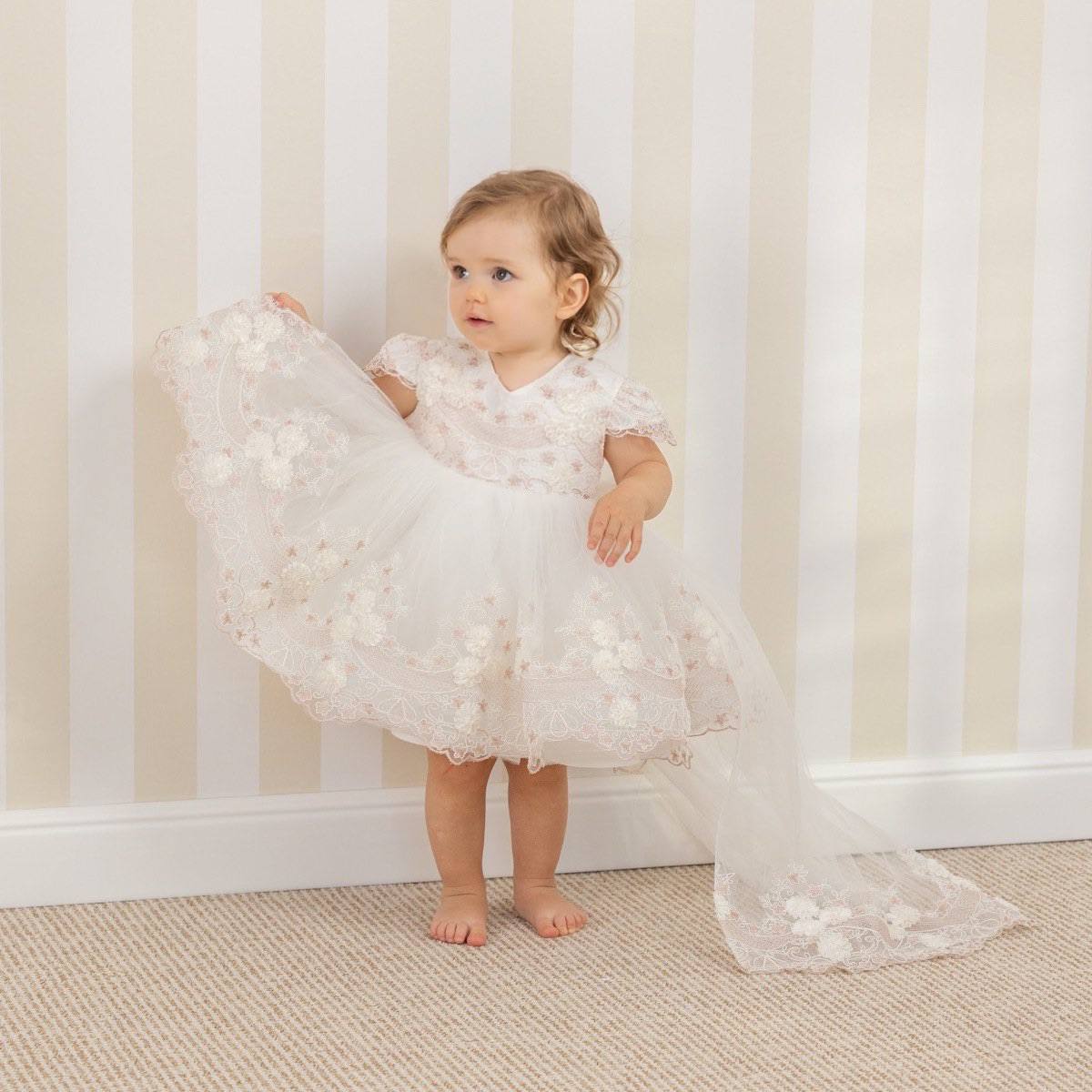 Baby in a  Baptism Dress with Detachable Train standing against a striped wall.