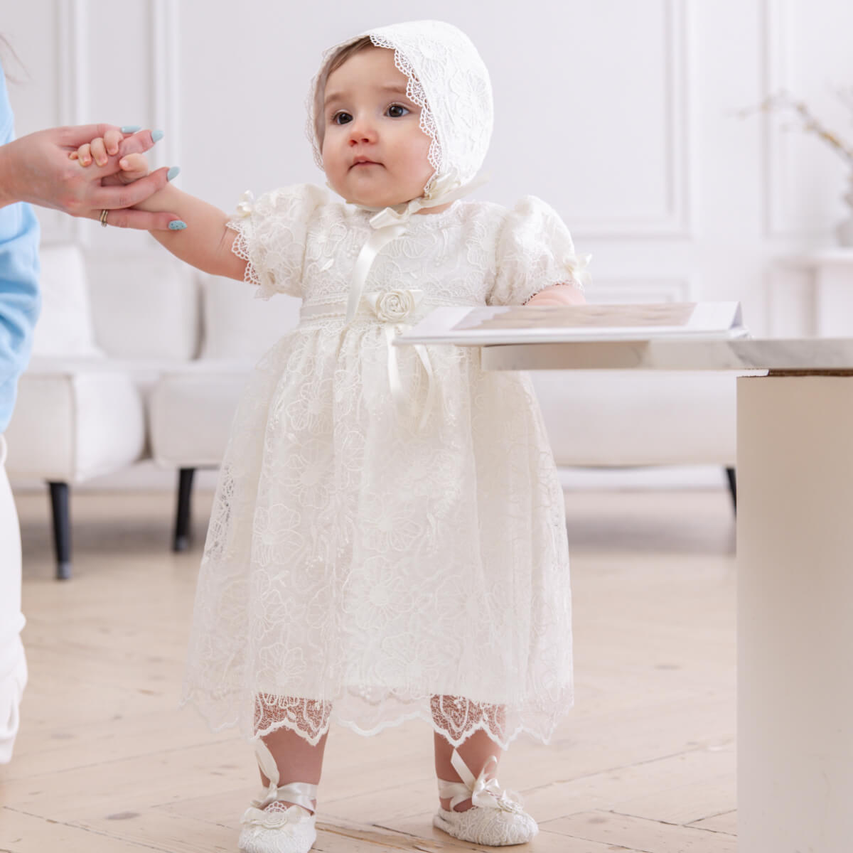 Baby in Christening Booties and bonnet standing on a light wooden floor.