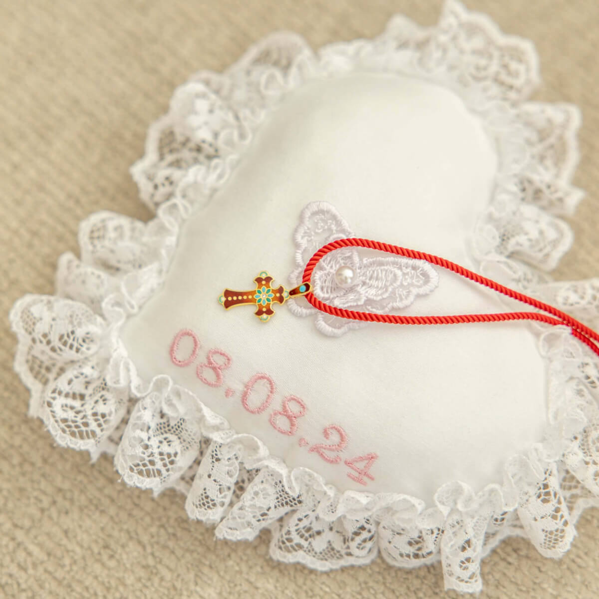Decorative heart-shaped pillow with lace trim, embroidered date, and a small cross pendant on a beige background.