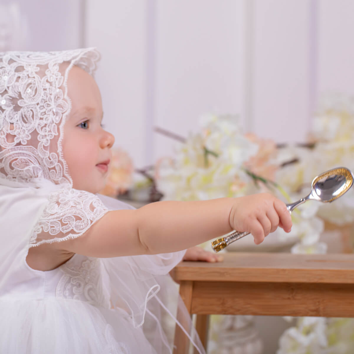 Child in a white lace outfit holding a spoon with a blurred floral background