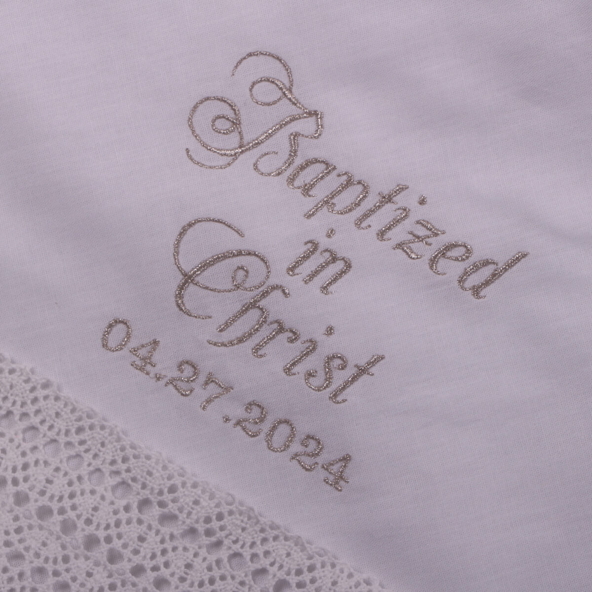 Embroidered text on a white fabric with lace edge, featuring 'Baptized in Christ' and a date.