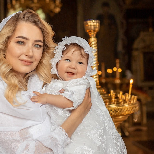 Woman holding a baby in a Lace Baptism Gown Set with Bonnet for Girl with a blurred background of candles and religious decor.