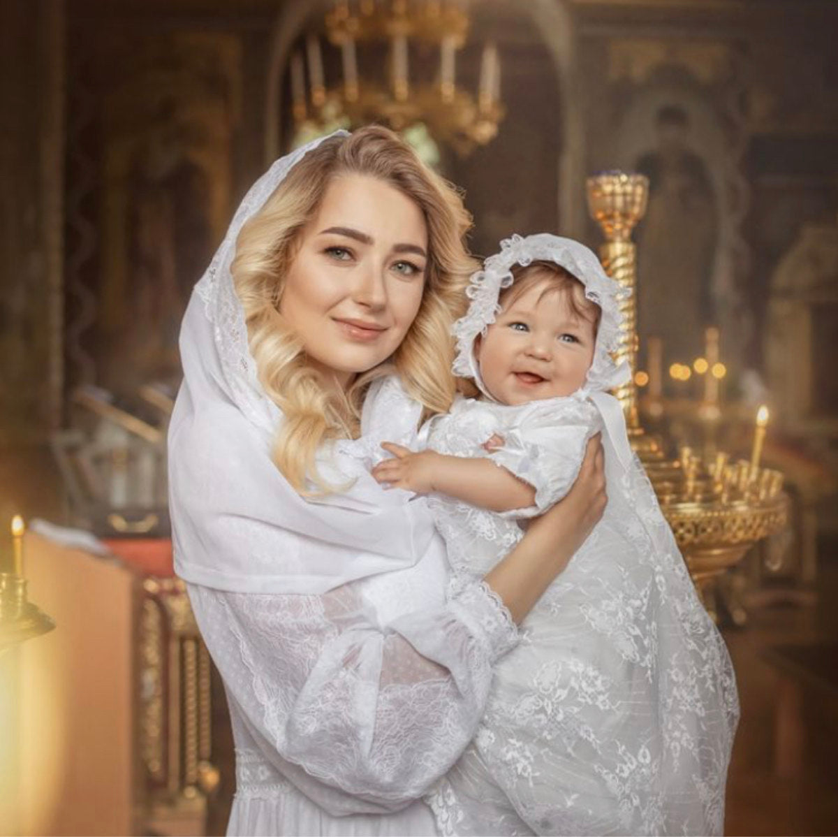 Woman holding a baby in a Lace Baptism Gown Set with Bonnet for Girl  with a blurred church interior background