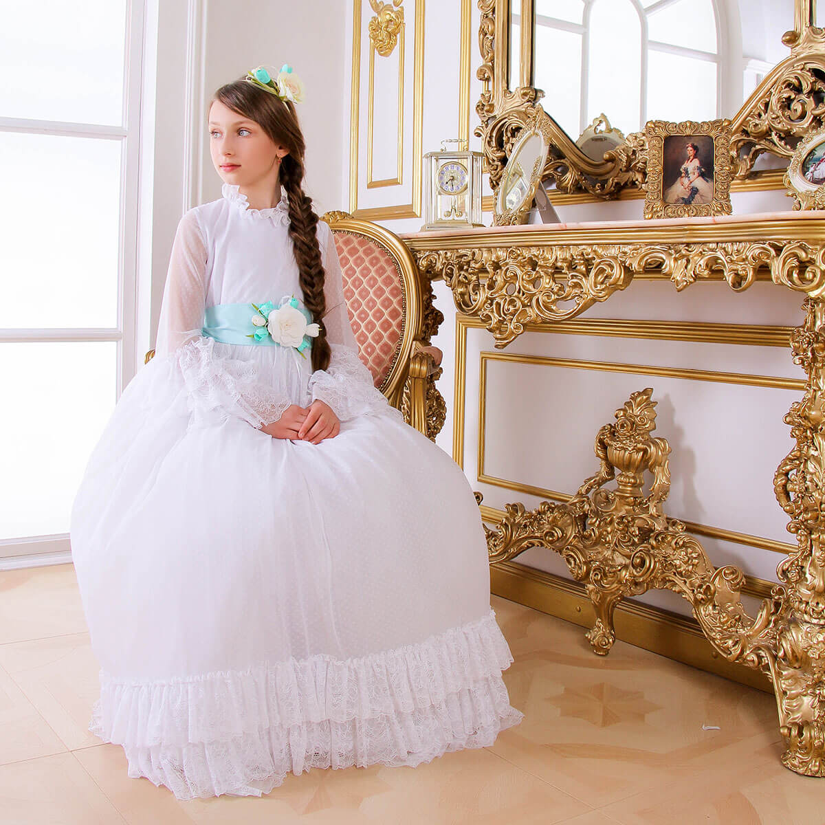 Young girl in Boho Chic Communion Dress sitting on an ornate gold chair in a bright room.
