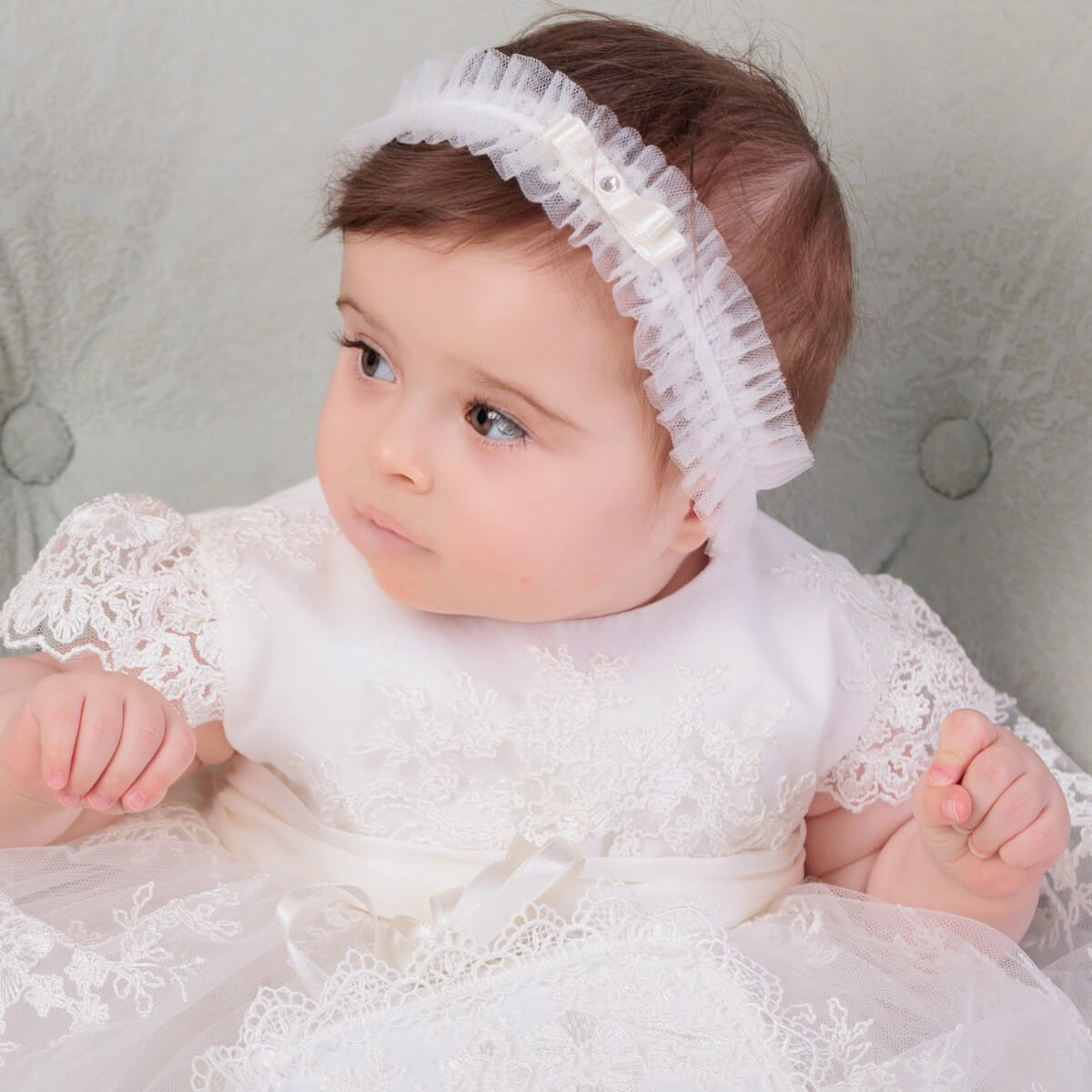 Baby in a white lace dress and baby headband against a neutral background