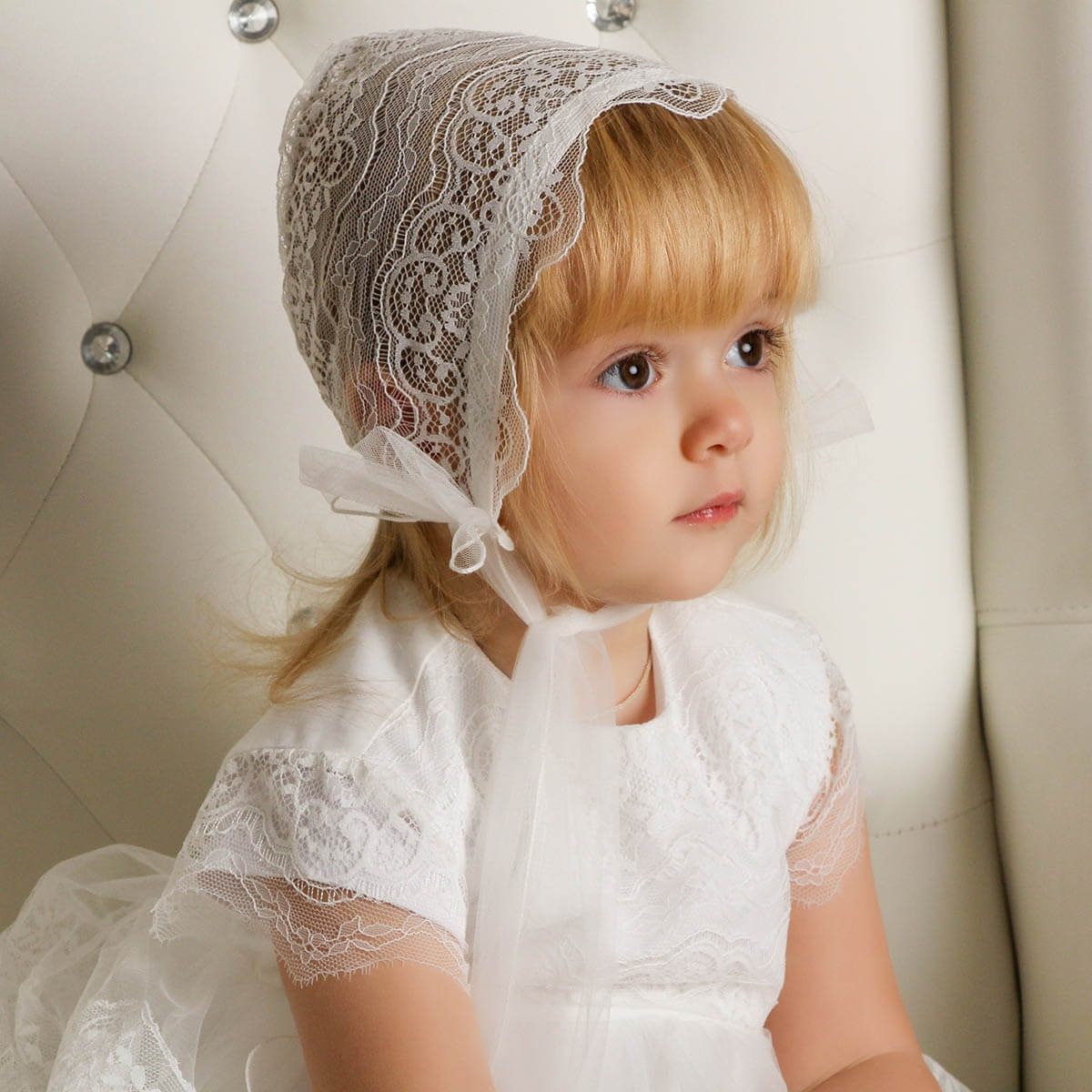 Young girl wearing a white lace dress and  Lace Baptism Bonnet sitting on a beige couch.