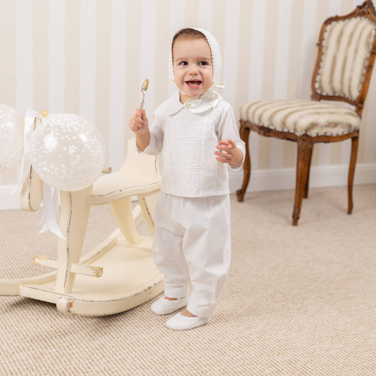 Child in a Baby Boy Baptism Suit standing in a room with a rocking chair and striped chair.