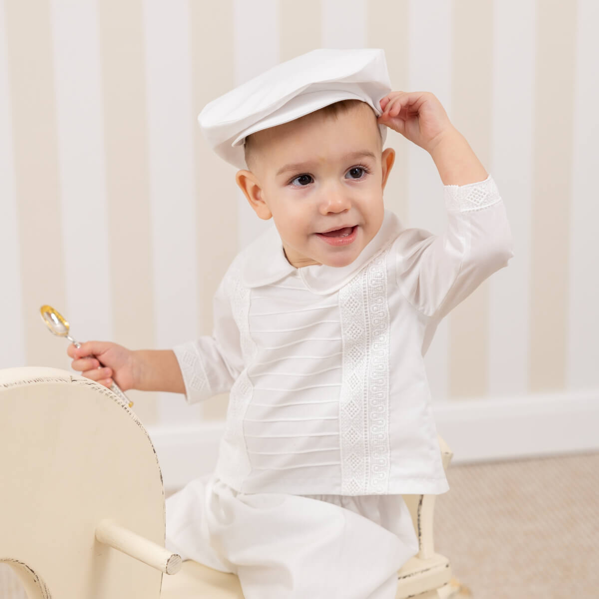 Child wearing a white outfit and Christening Cap, sitting on a chair with a neutral background