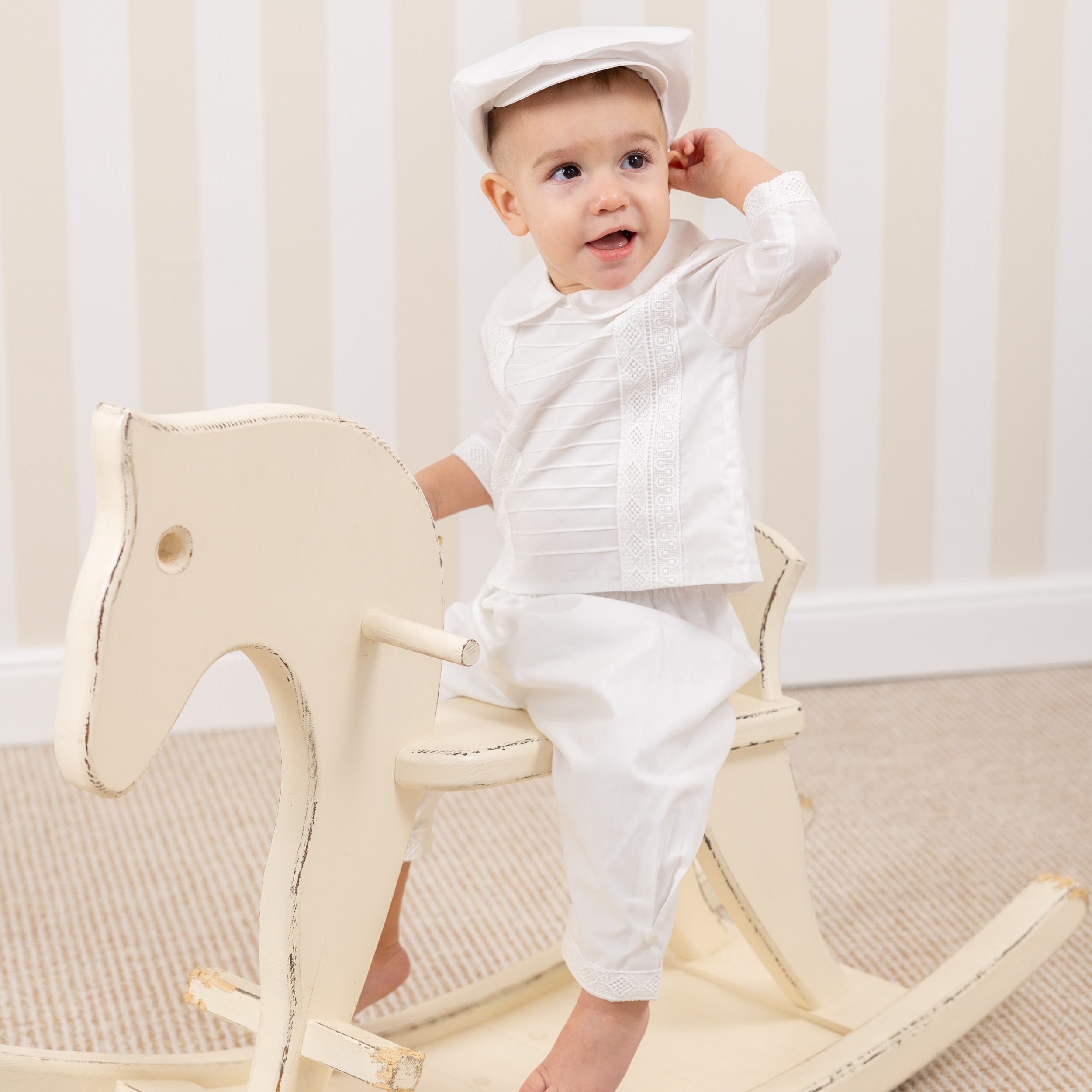 Baby in a Christening Cap sitting on a wooden rocking horse against a neutral background
