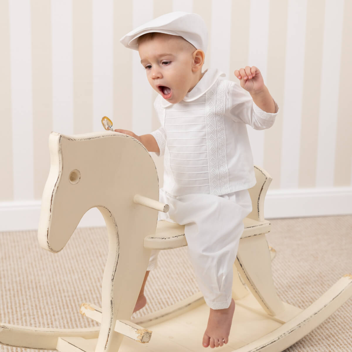 Child in a Christening Cap playing on a wooden rocking horse in a room with white curtains.