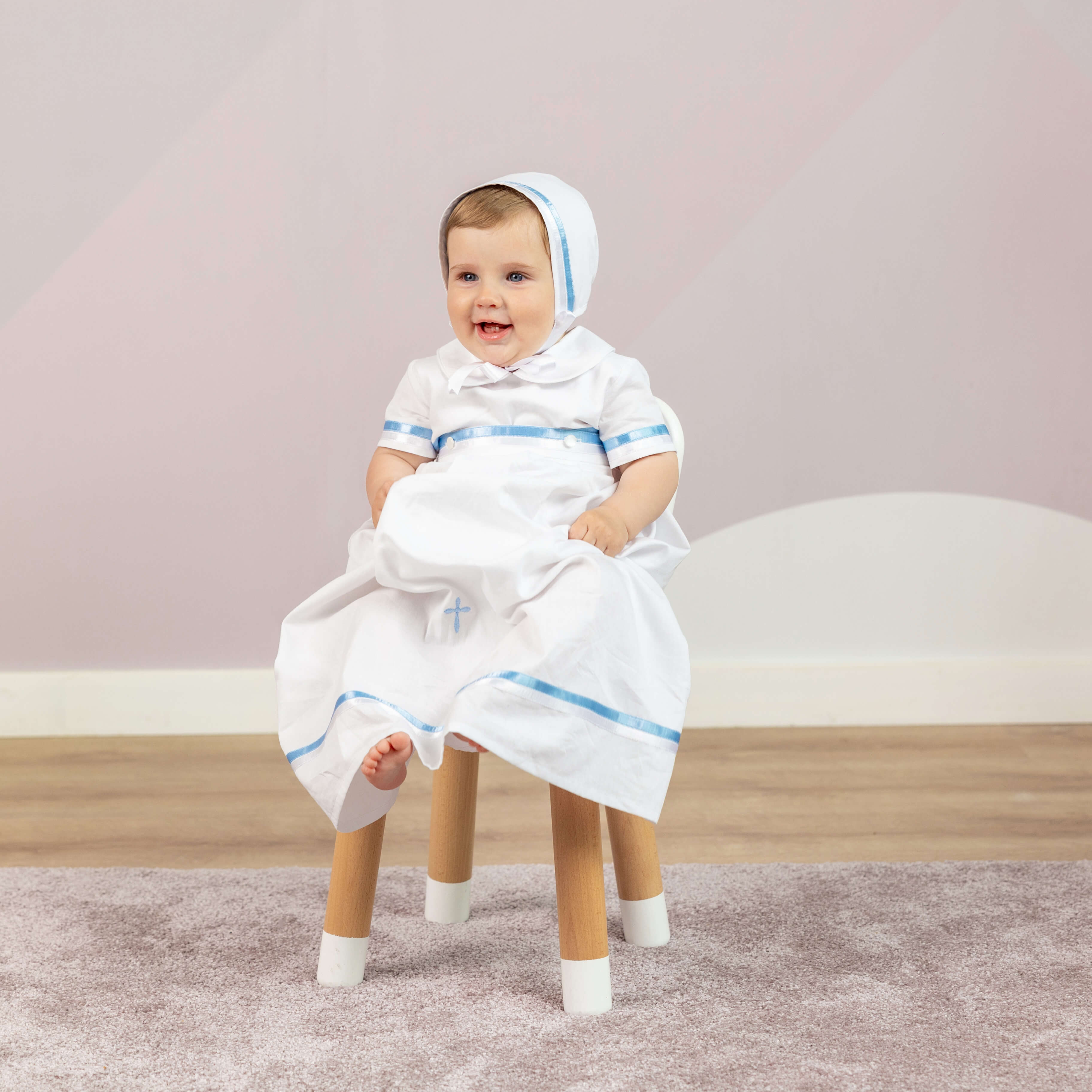 Baby in a white white christening gown with blue trim sitting on a stool against a light pink wall.