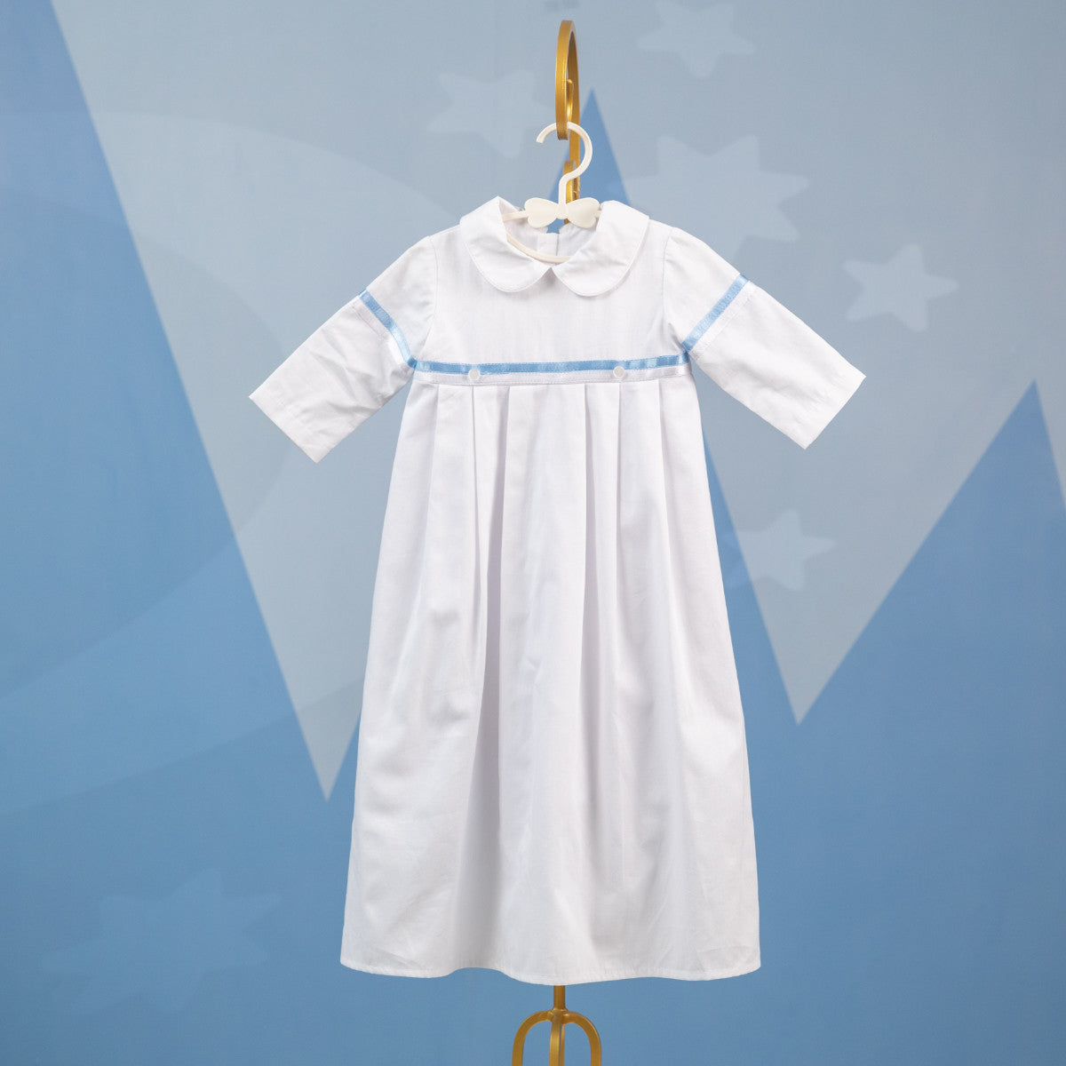 white christening gown with blue trim on a hanger against a blue geometric background