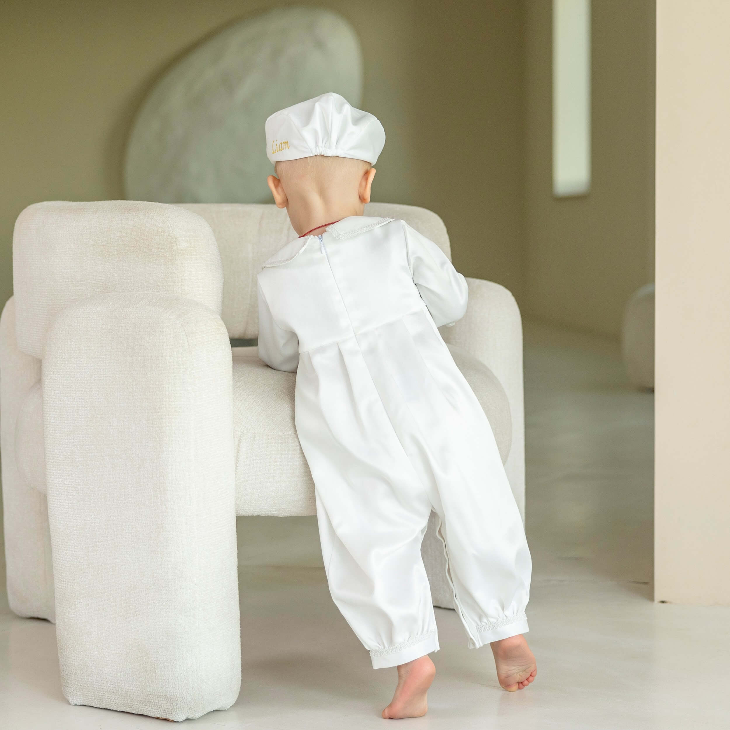Baby in a Boy Christening Outfit standing next to a white chair in a minimalistic room.