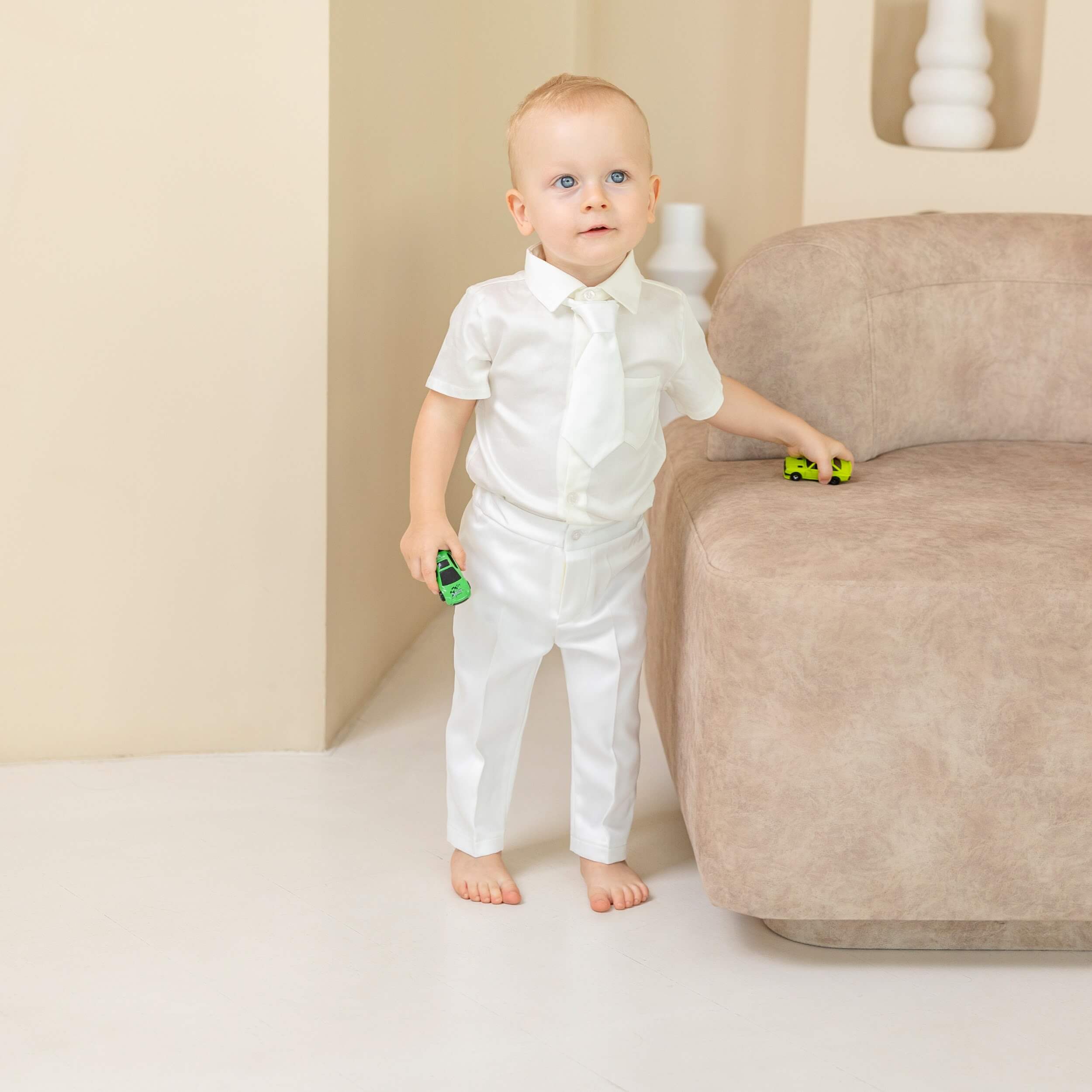 Baby in a Baby Boy Baptism Suit standing next to a beige sofa holding toy cars.