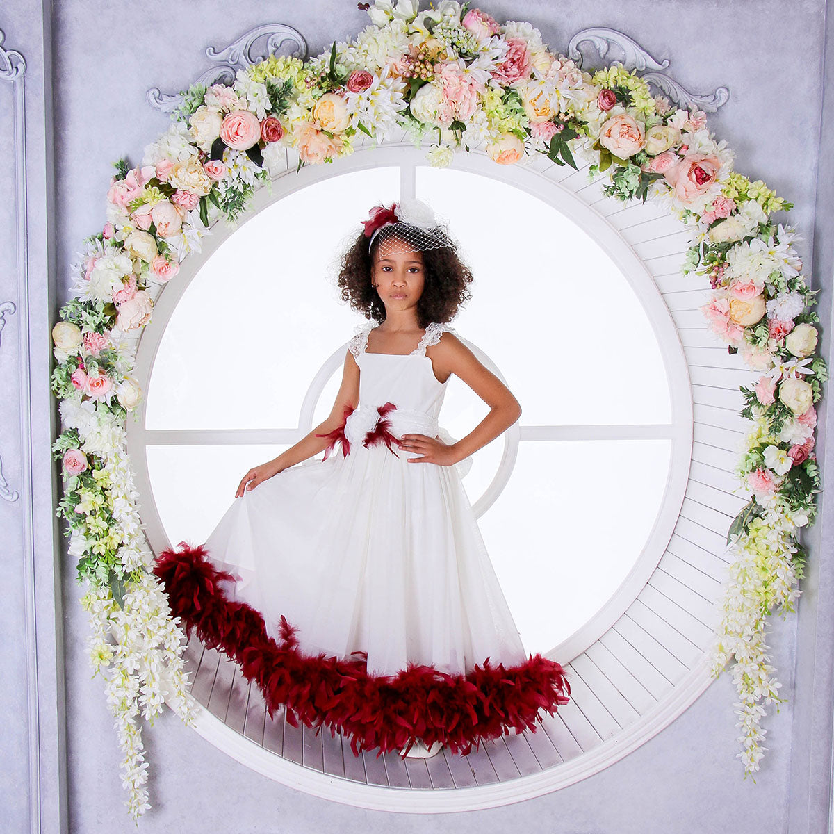 Young girl in a white dress with red feathered skirt standing in front of a floral arch.