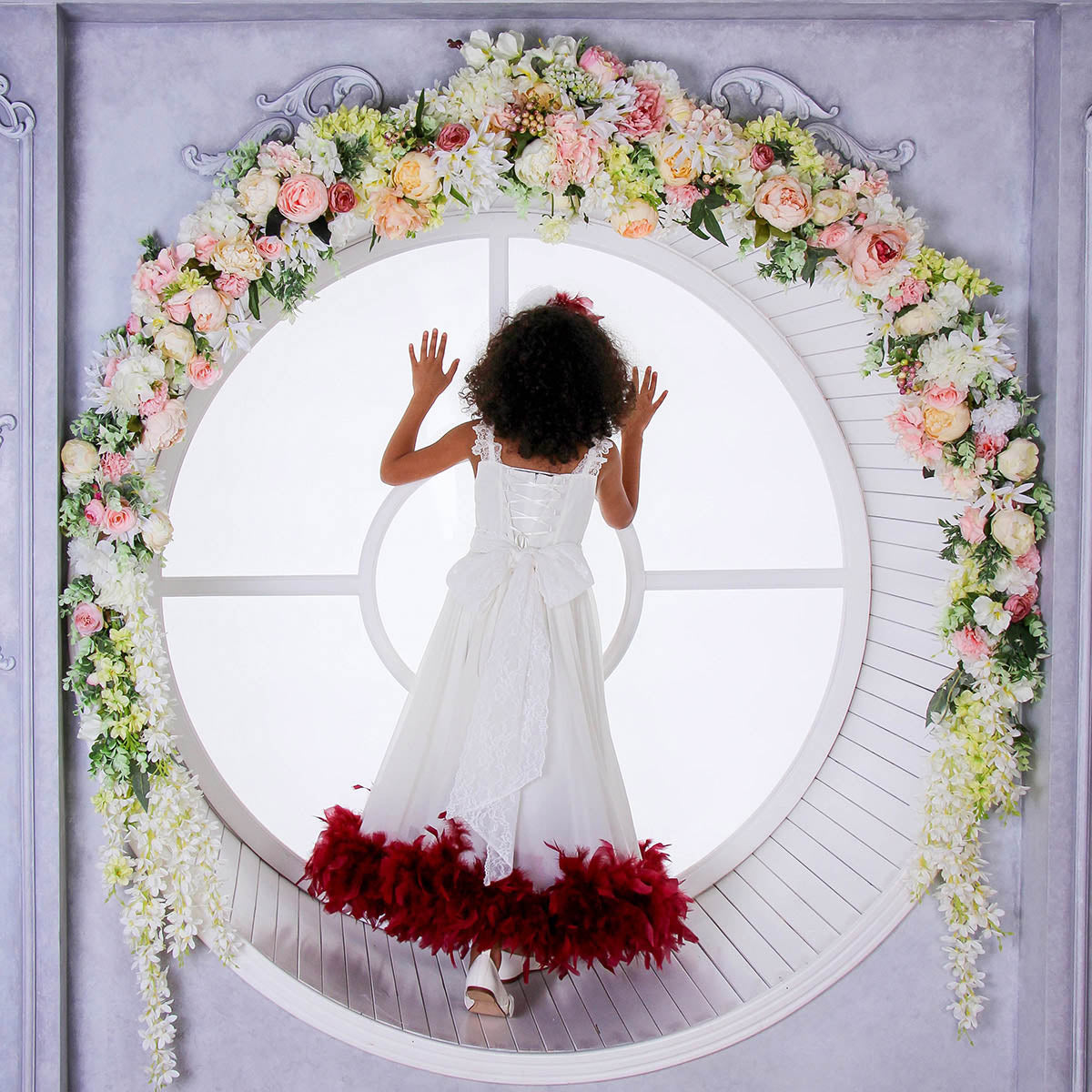 Child in a white dress with red feathered skirt standing in front of a floral arch.