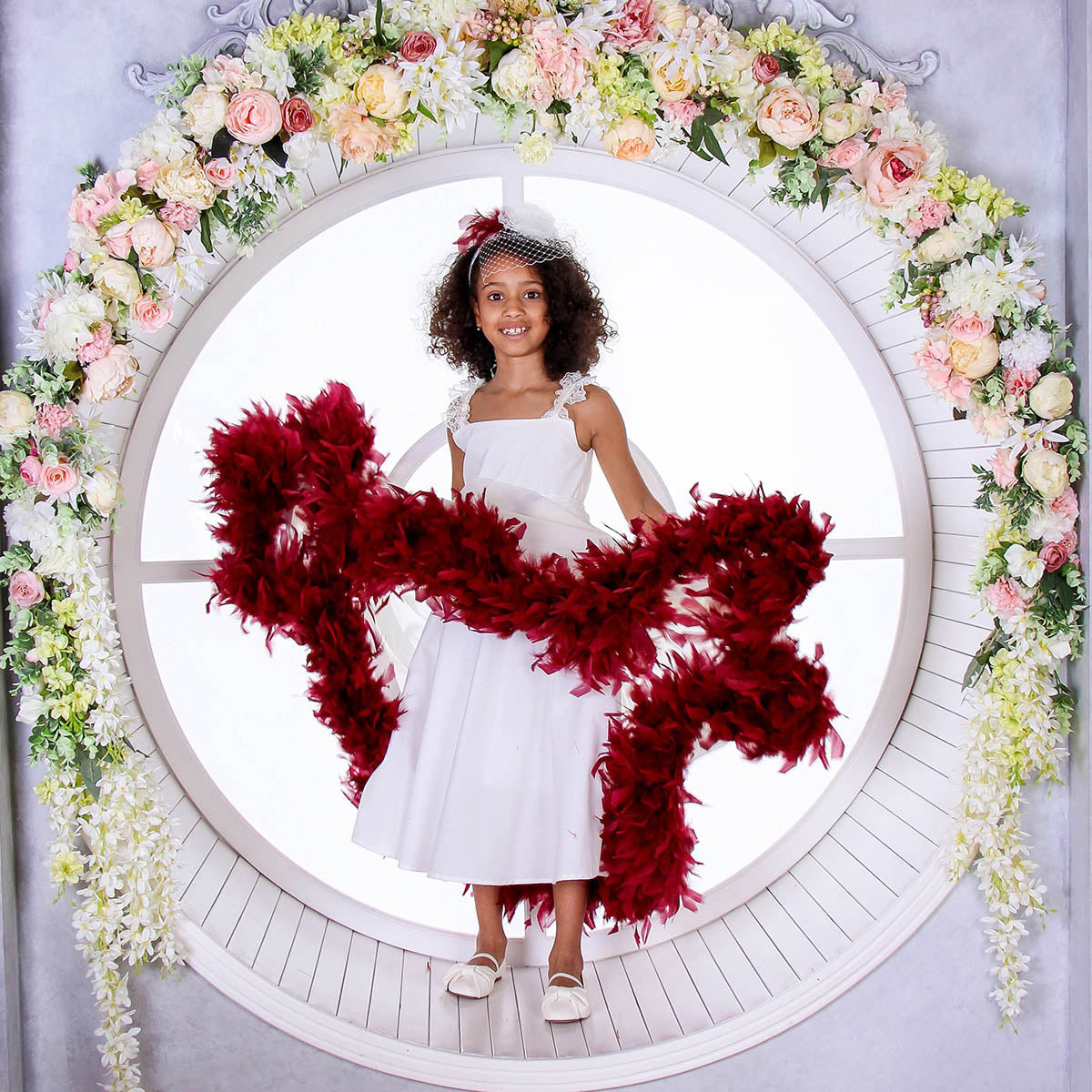 Young girl in a white dress with red feathered wings standing in front of a floral wreath.