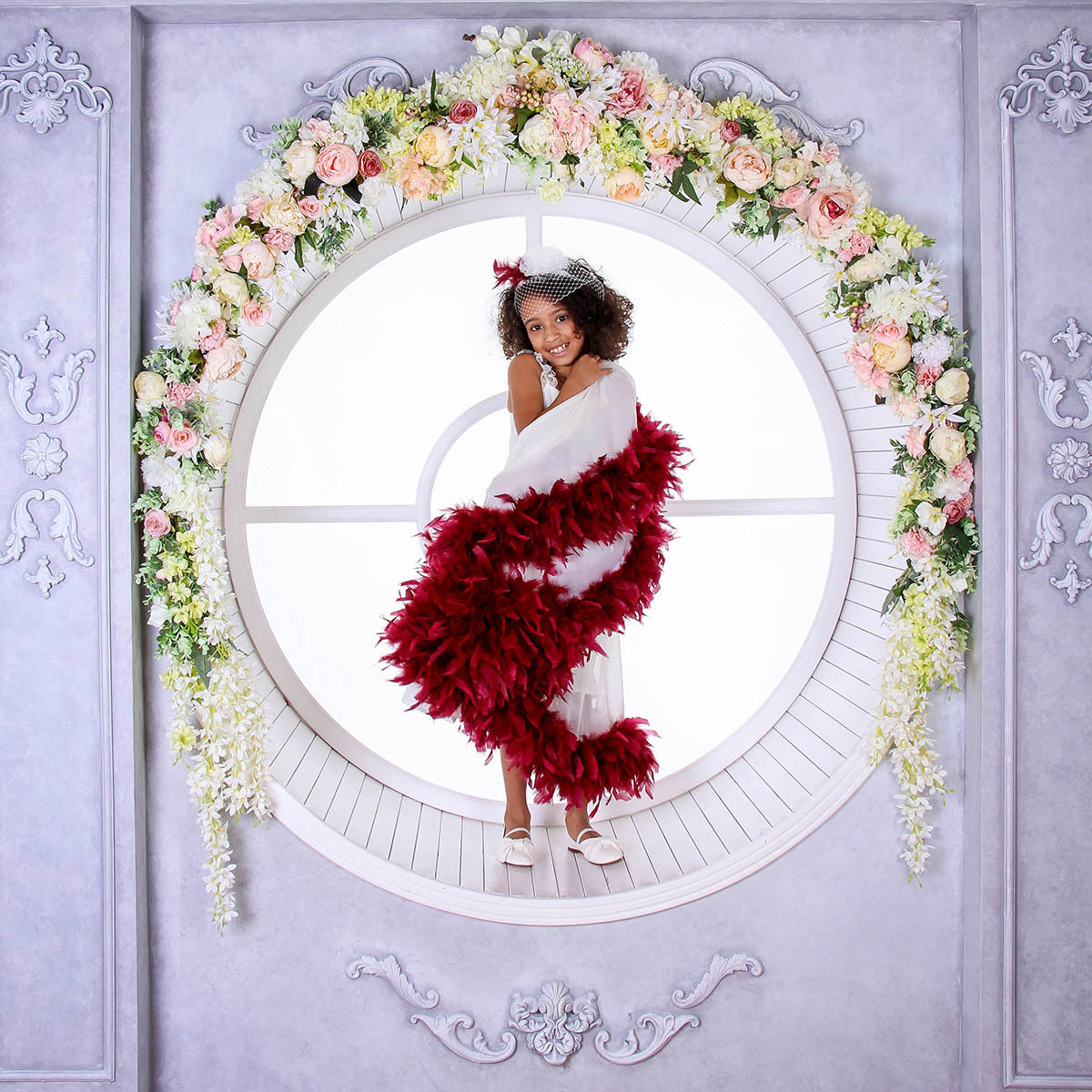 Young girl in a red and white dress with feather boa standing in front of a decorative floral frame.