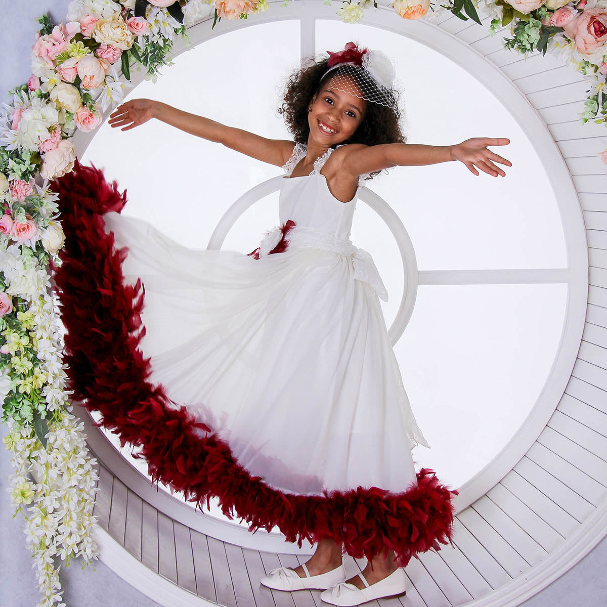 Young girl in a white dress with red feathered skirt standing in a circular frame surrounded by flowers.