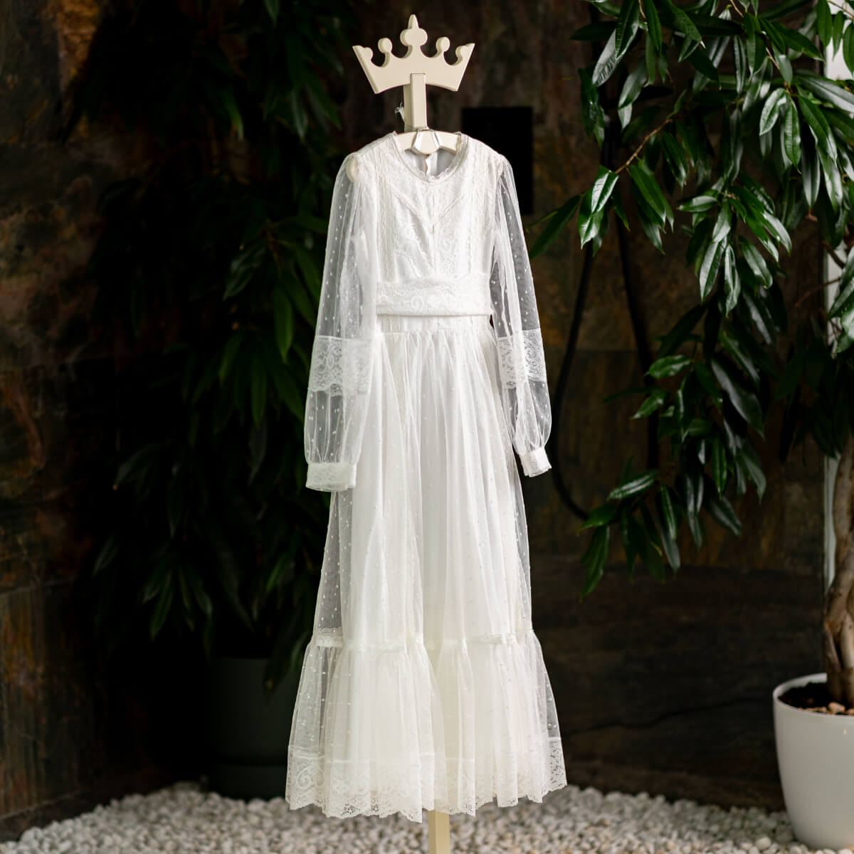 White lace Traditional Communion Dress on a mannequin against a dark background with plants.
