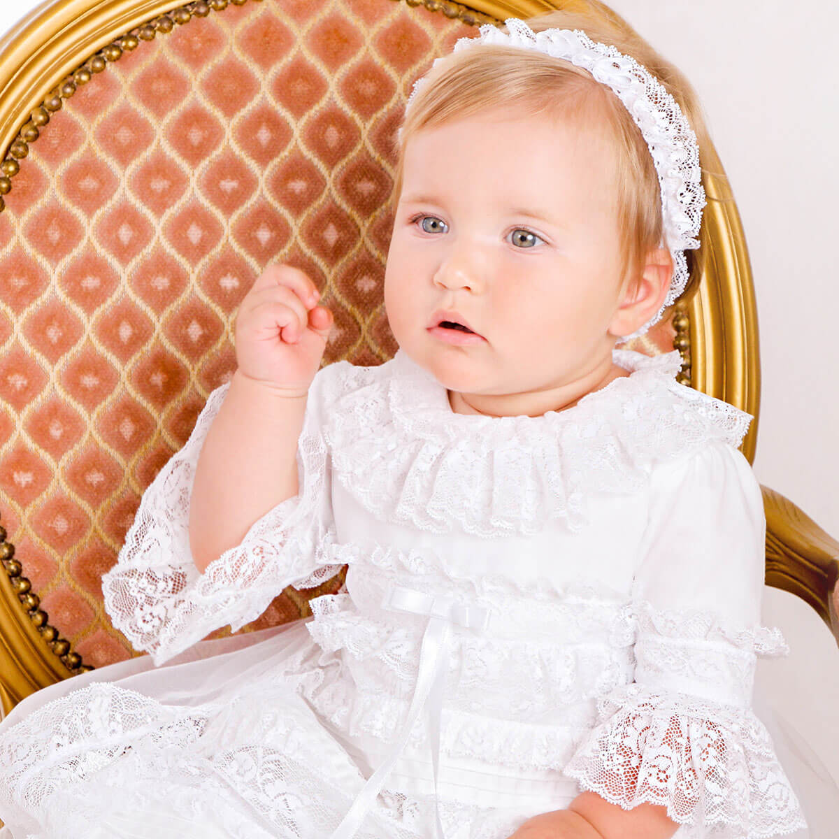 Baby in a white lace dress sitting on a decorative chair.