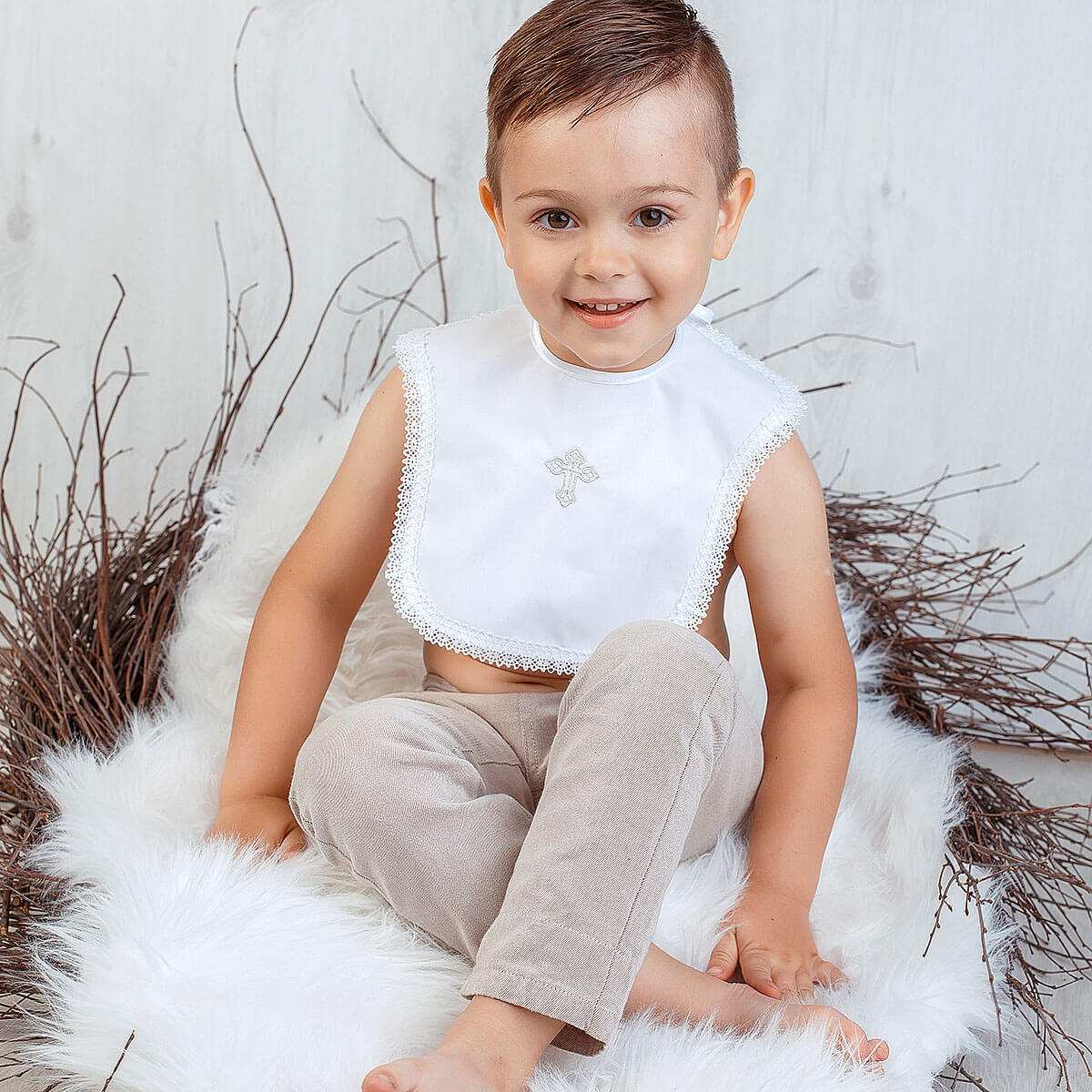 Child wearing a Baptism Bib with Cross Embroidery with lace trim sitting on a fluffy white surface with decorative branches.