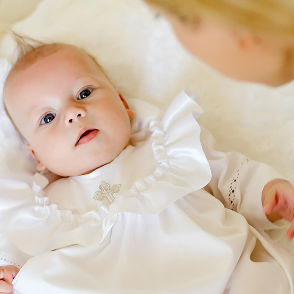 Baby in a christening long gown lying on a white blanket with another person partially visible.