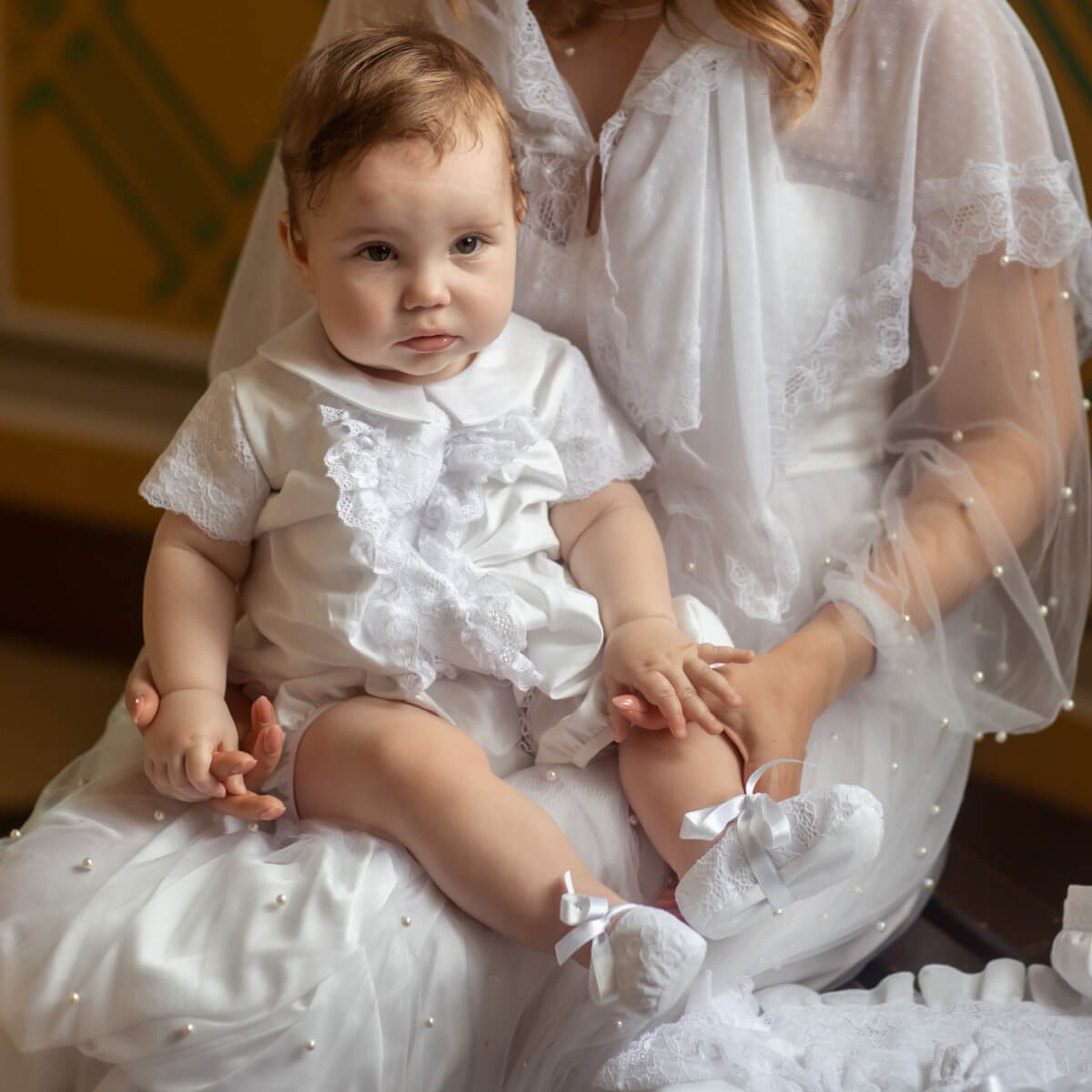 Baby in a white baptism outfit sitting on a person in a white dress with lace details.