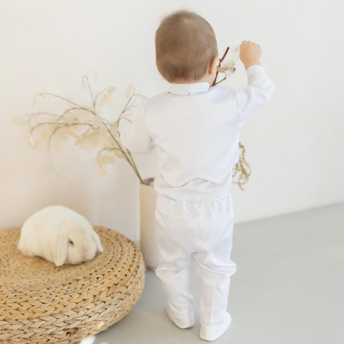 Child in a  Baby Boy Baptism Suit standing next to a small white rabbit on a woven mat.