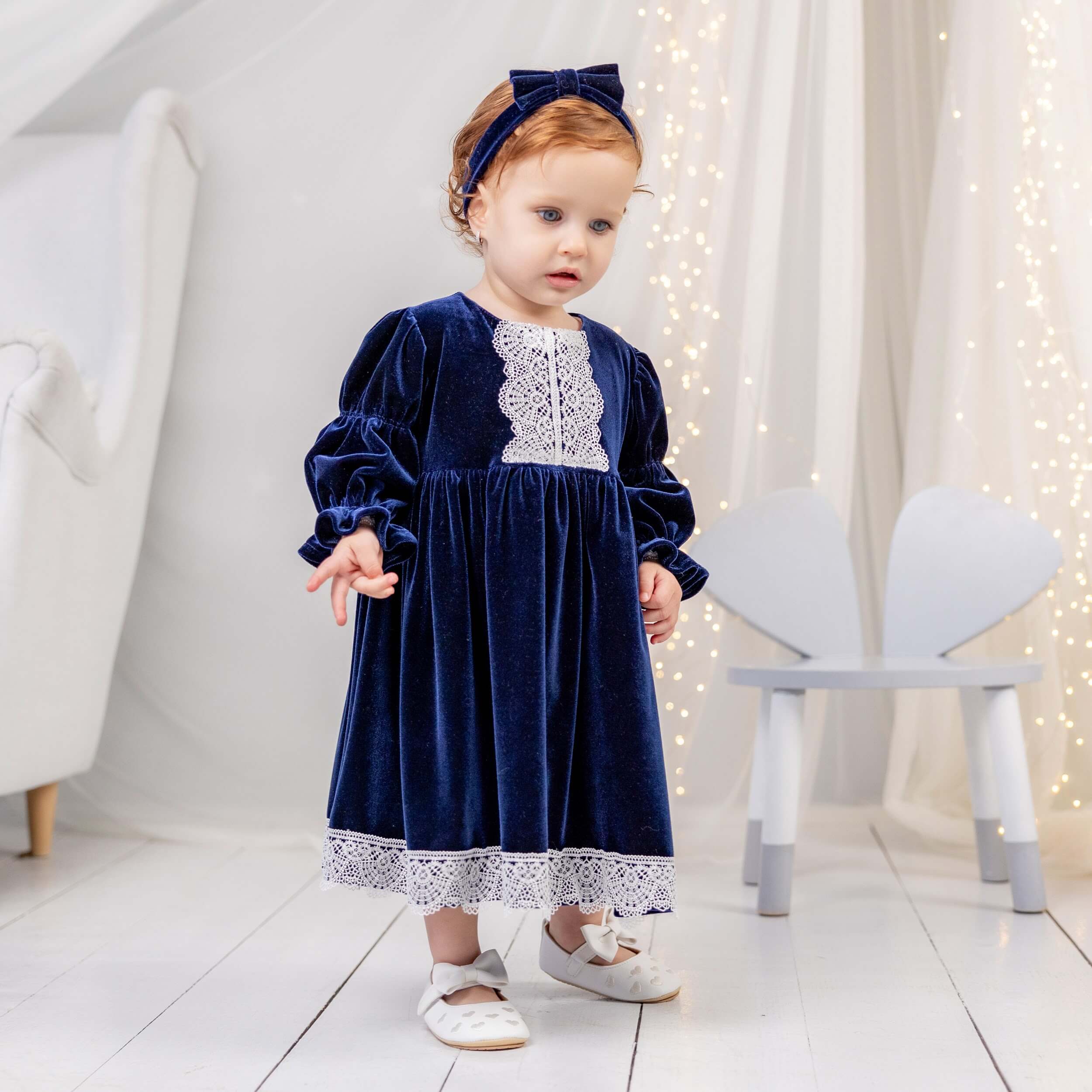Young child wearing a navy blue velvet dress with lace details in a softly lit room.